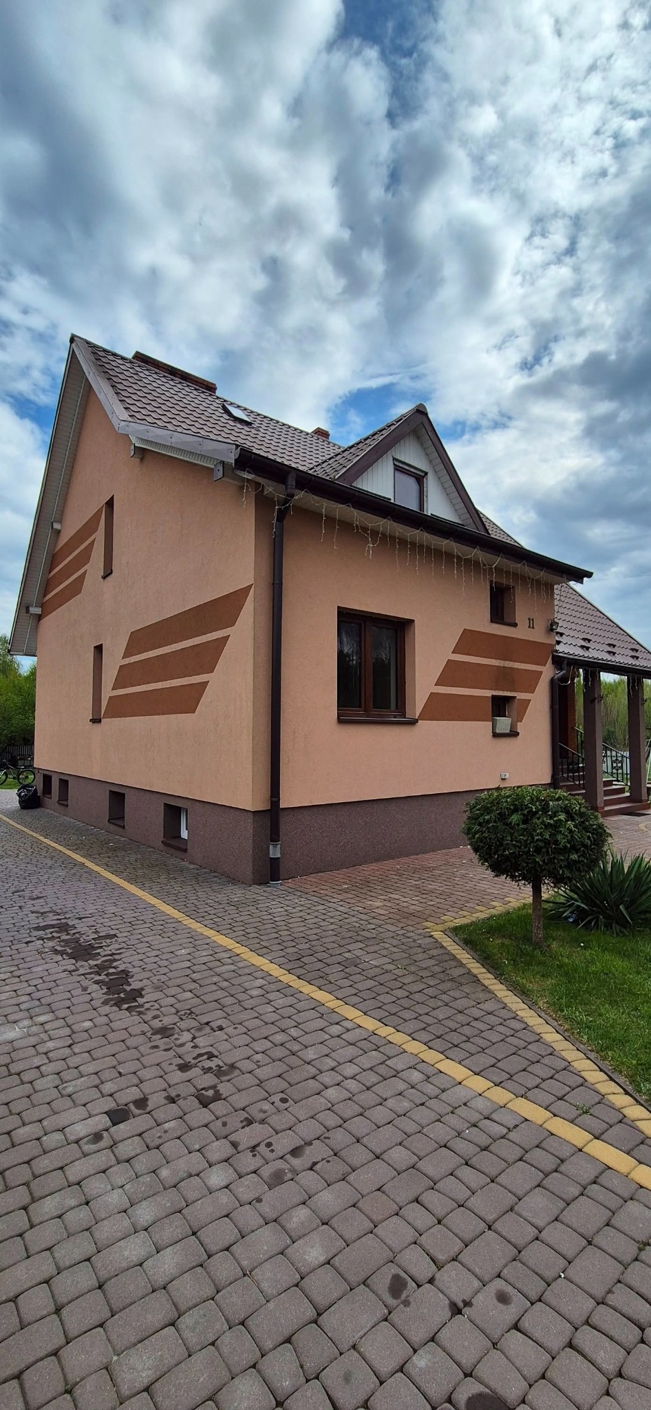 Dom z brązowymi oknami PCV w Radomiu, widok z zewnątrz. Dwukondygnacyjny budynek z dekoracyjnymi pasami i brązową podmurówką. Brukowany podjazd.