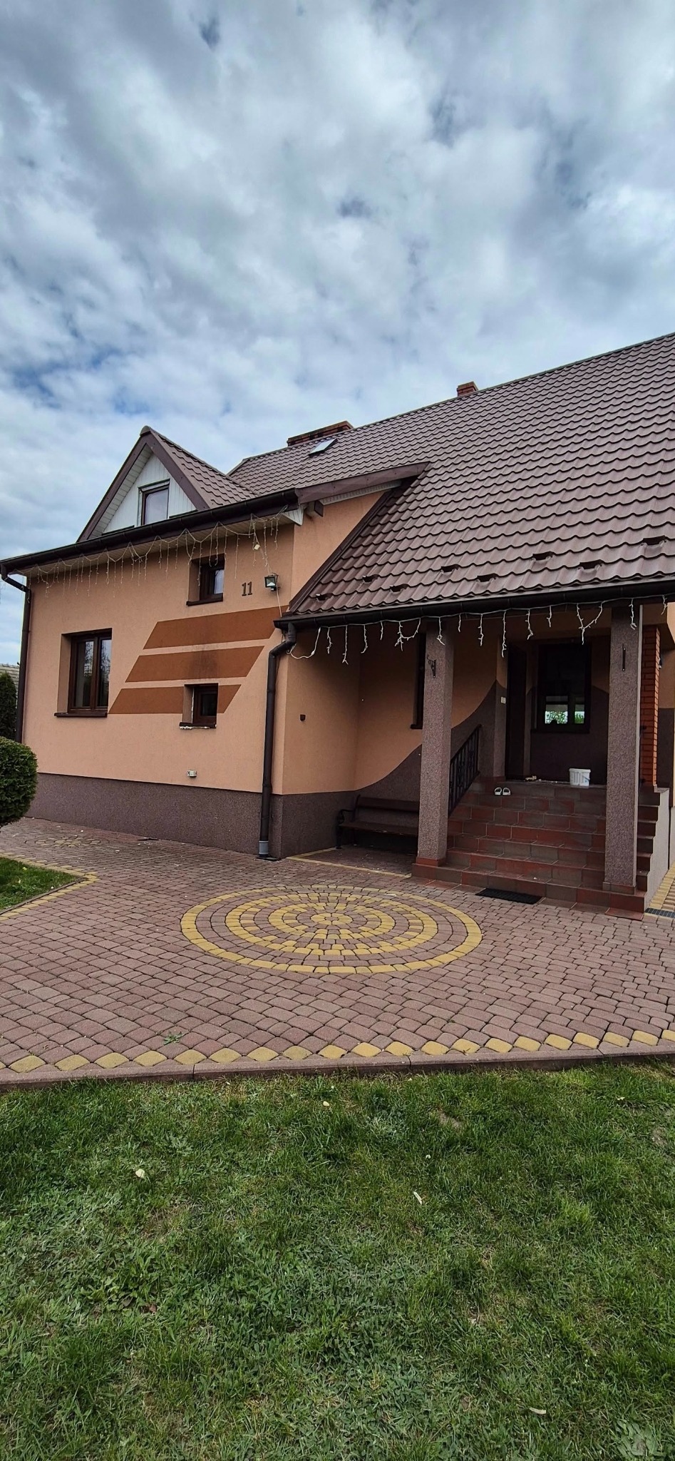 Dom z brązowymi oknami, beżową elewacją i dekoracyjną kostką brukową. Widoczne okna z brązowymi ramami, dopasowane do koloru dachu i elementów wykończeniowych.