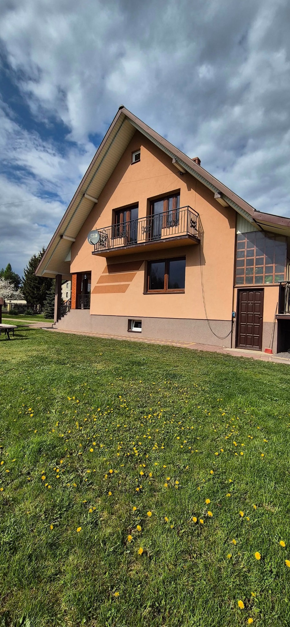 Dwupiętrowy dom z balkonem i brązowymi oknami, otoczony trawnikiem z żółtymi kwiatami. Widoczne elementy architektoniczne i detale wykończeniowe.