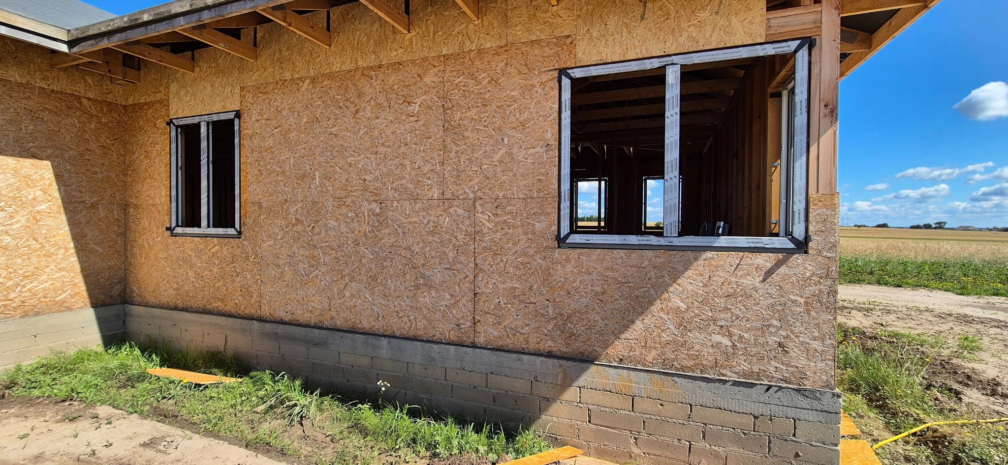 Montaż okien w domu w trakcie budowy. Widoczne ramy okienne w ścianie z płyt OSB. W tle pole i błękitne niebo. Okna marki Salamander.