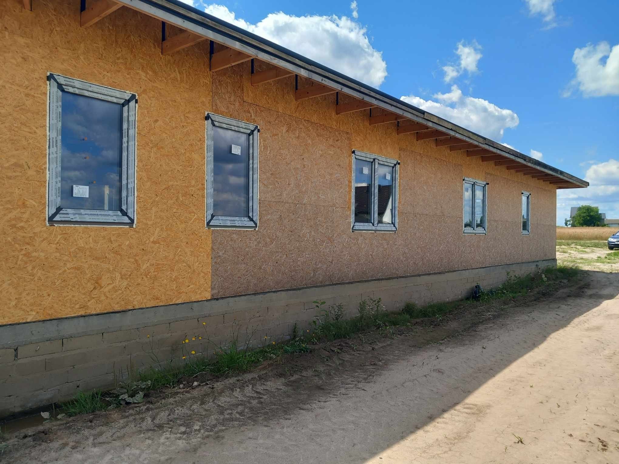 Montaż okien w domu szkieletowym, widoczne okna z folią ochronną i płyty OSB na elewacji. Budynek w trakcie budowy, w tle błękitne niebo.