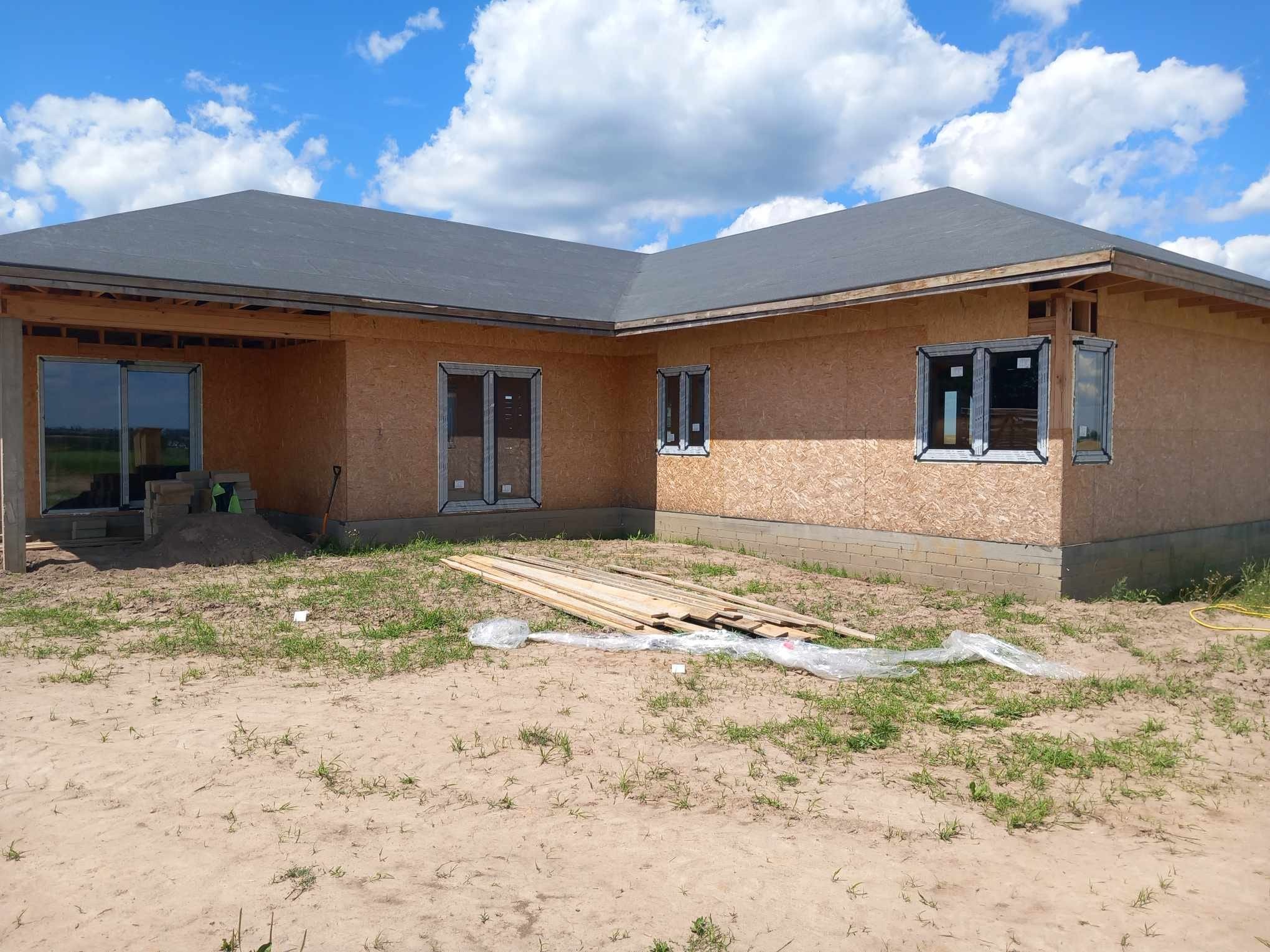 Dom w trakcie budowy z zamontowanymi oknami. Widoczne płyty OSB na ścianach, szare pokrycie dachu i ułożone deski na piaszczystym terenie.