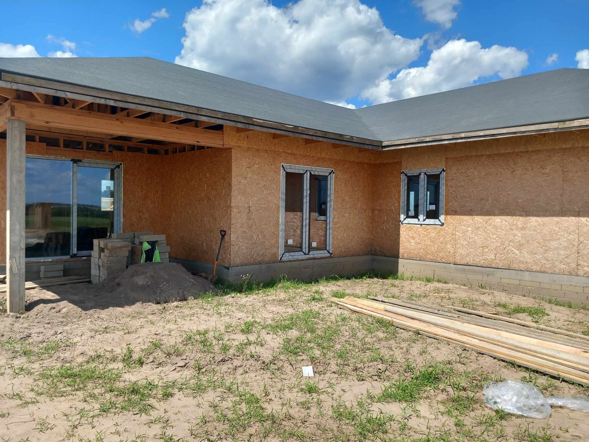 Montaż okien w surowym domu z płyt OSB, widoczne ramy okienne i przygotowane otwory. Na pierwszym planie piasek, cegły i deski. Błękitne niebo z chmurami w tle.