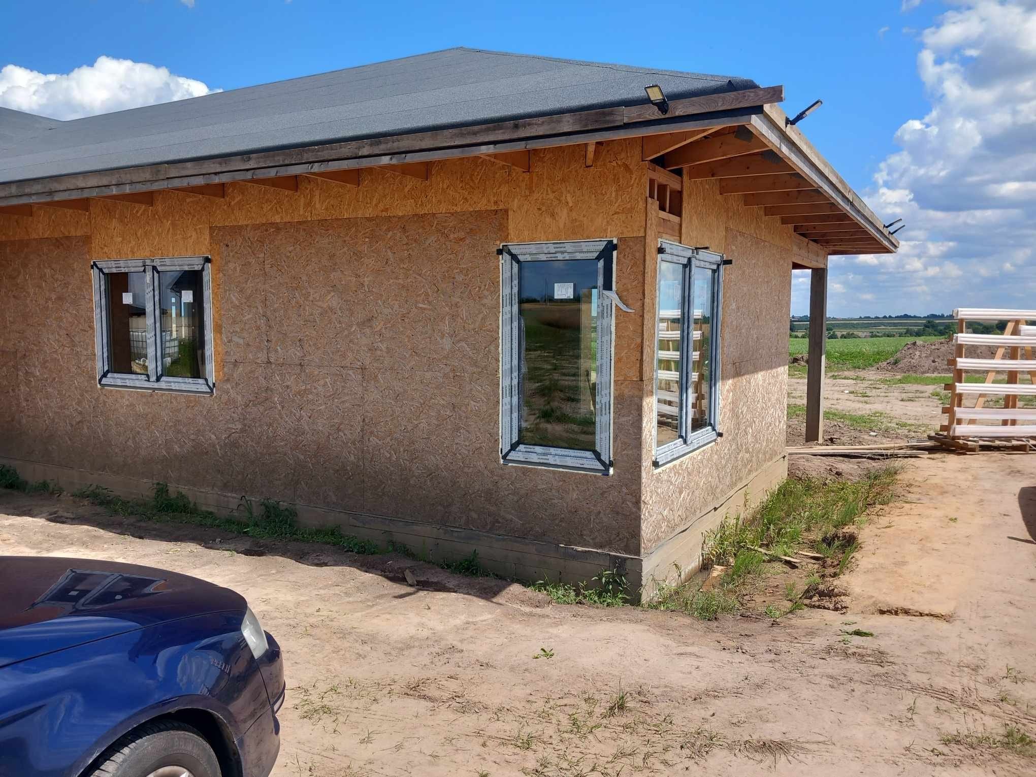 Dom w trakcie budowy, z zamontowanymi białymi oknami w ścianach z płyty OSB. W tle widoczne pola i niebo.