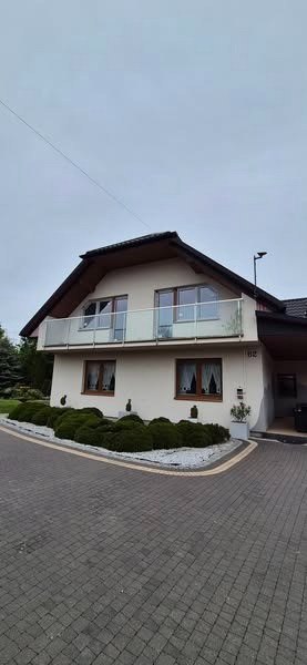 Dwupiętrowy dom z balkonem z przeszkloną balustradą i brązowymi ramami okiennymi, otoczony zielenią i kostką brukową.