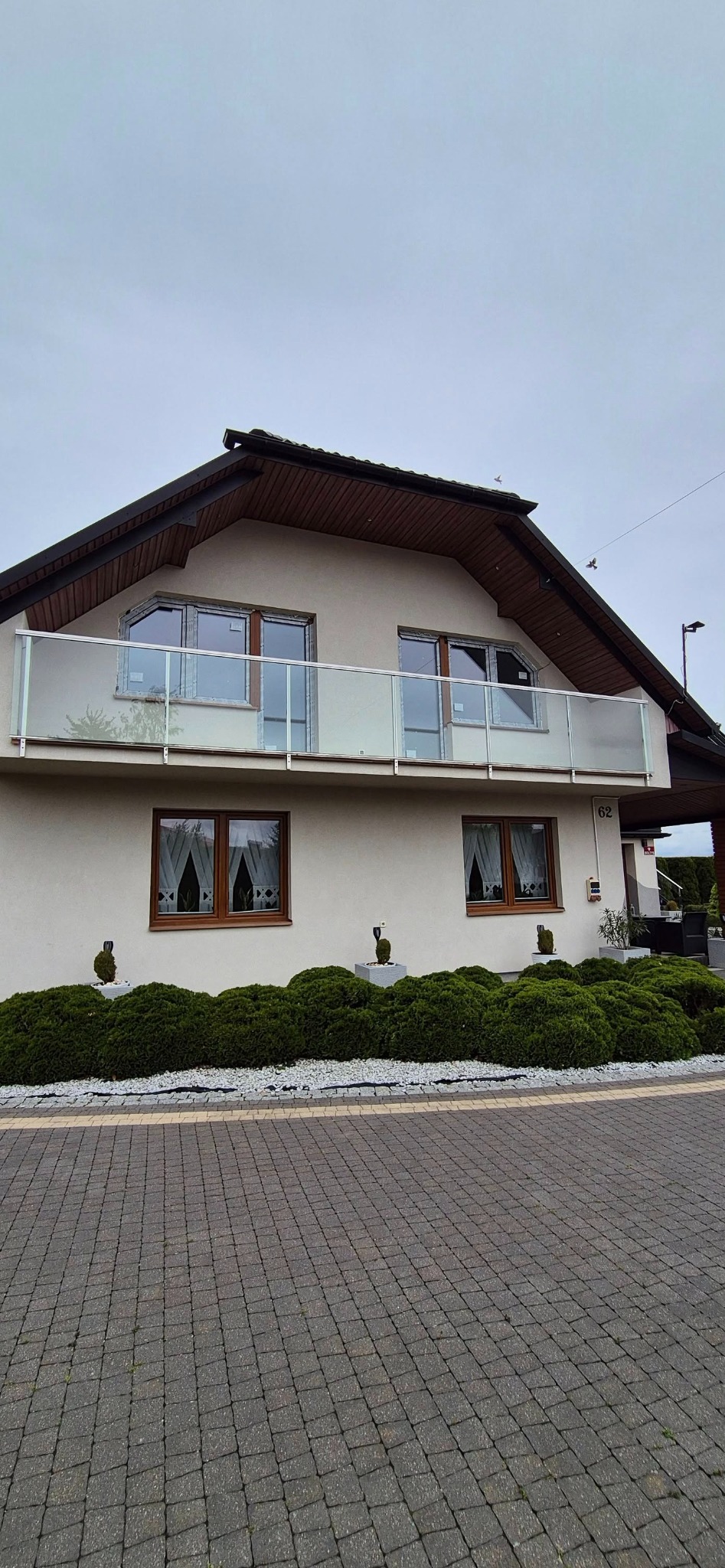 Dom z balkonem z przeszkloną balustradą i oknami z drewnianymi ramami, otoczony zielenią i brukowaną ścieżką. Na ścianie widoczny numer domu.