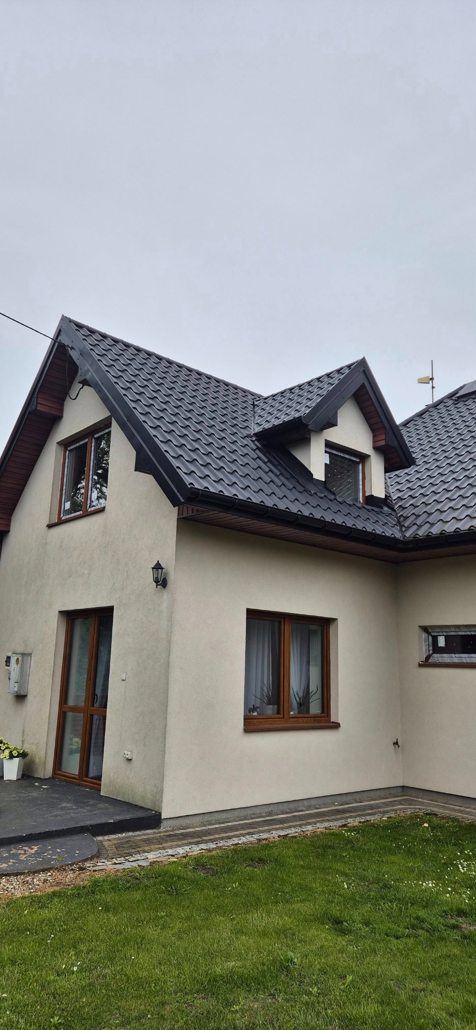 Dom z beżową elewacją i brązowymi oknami PCV, czarny dach z dachówki, widok z dołu, pochmurne niebo. Zewnętrzna lampa ścienna.