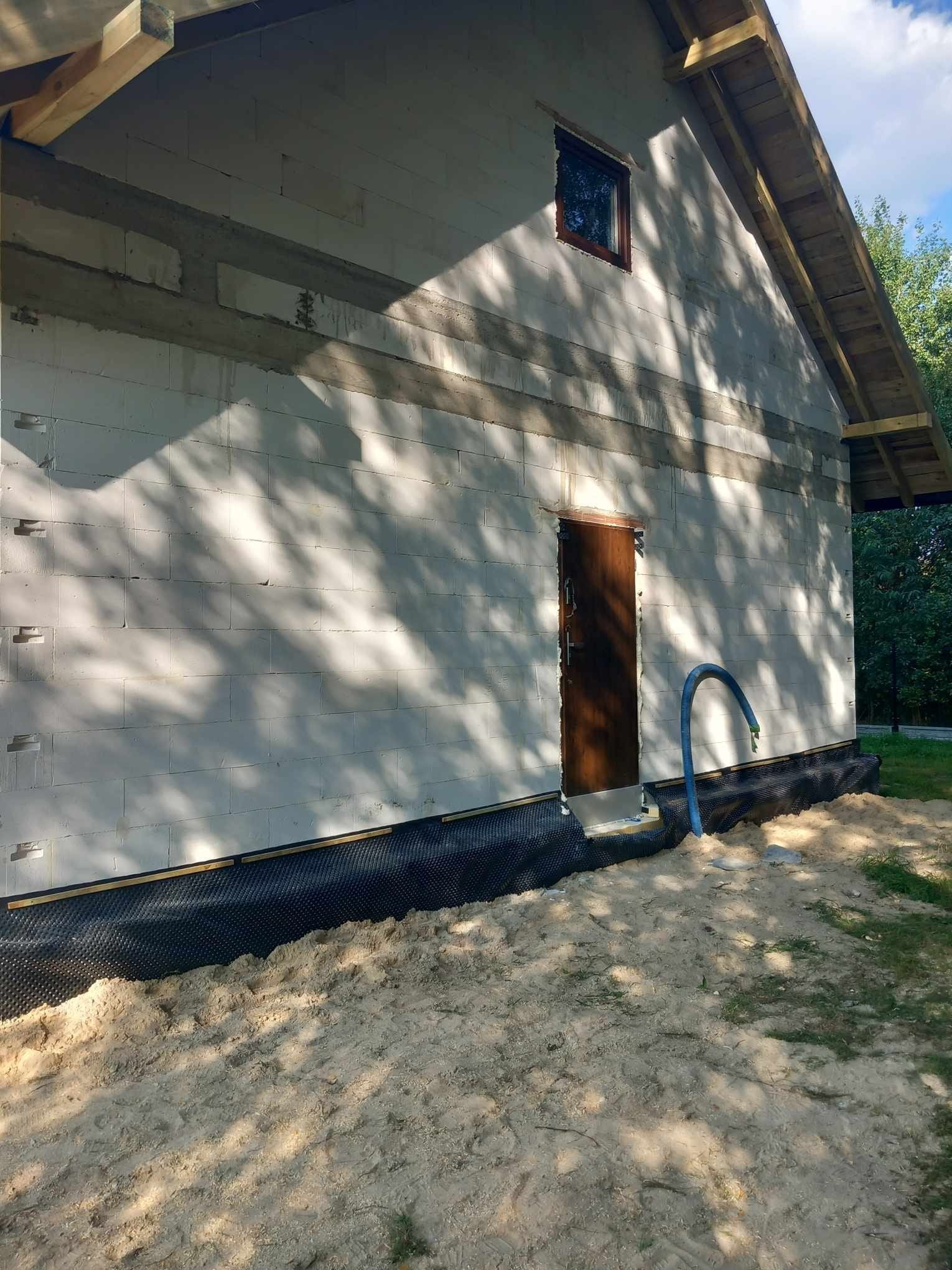 Budowa domu z pustaków: widoczna ściana z oknem w ramie, drzwi wejściowe i fragment dachu. W tle drzewa, na pierwszym planie piasek.