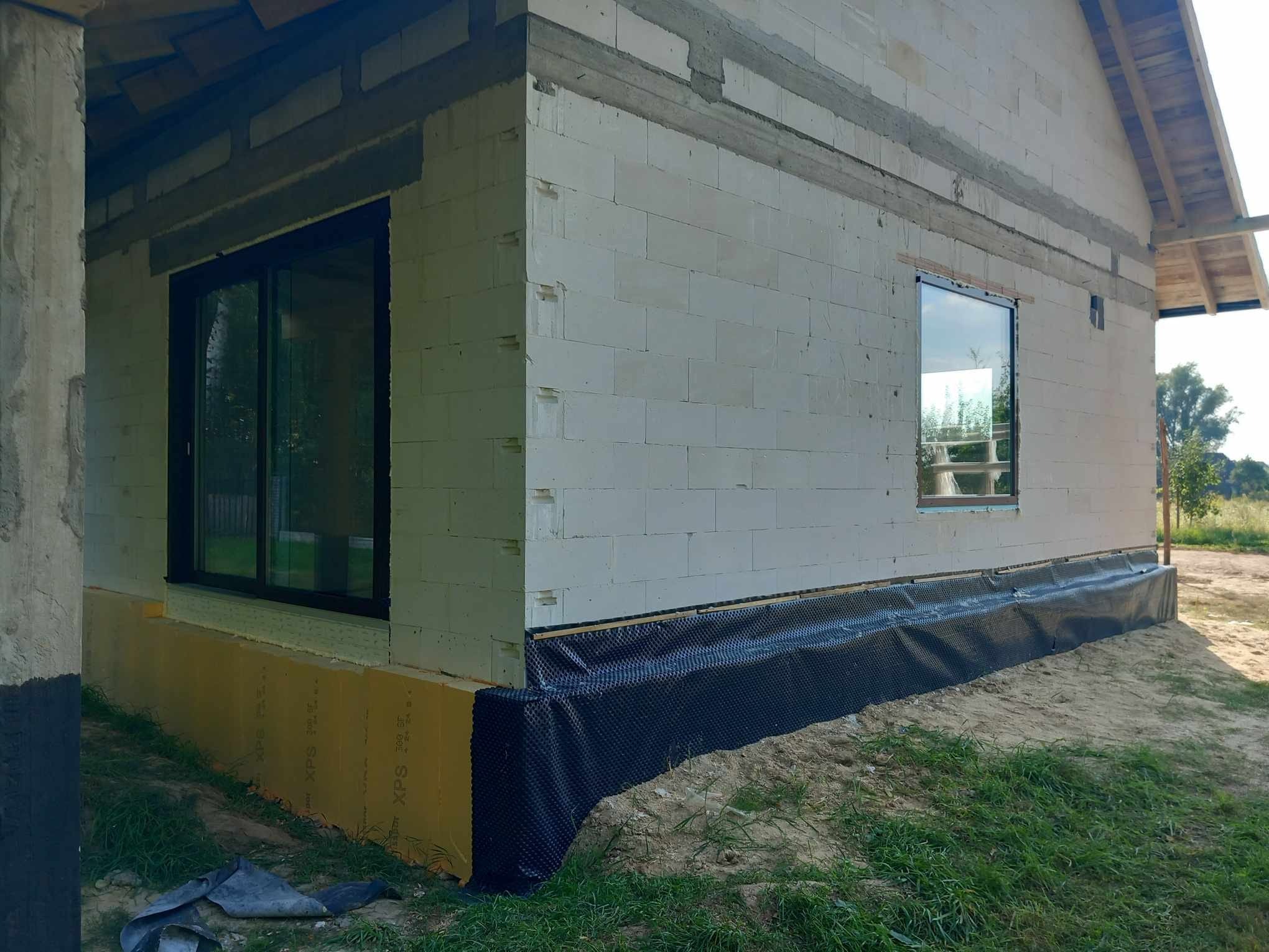 Montaż okien w surowym domu z białego betonu komórkowego. Widoczne czarne okna i izolacja fundamentów. Dom w trakcie budowy, otoczenie: trawa, piasek.