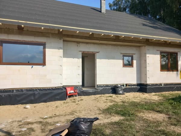 Montaż okien z brązową ramą w surowym domu z białego bloczka, widoczne ocieplenie fundamentów i przygotowanie do dalszych prac wykończeniowych.