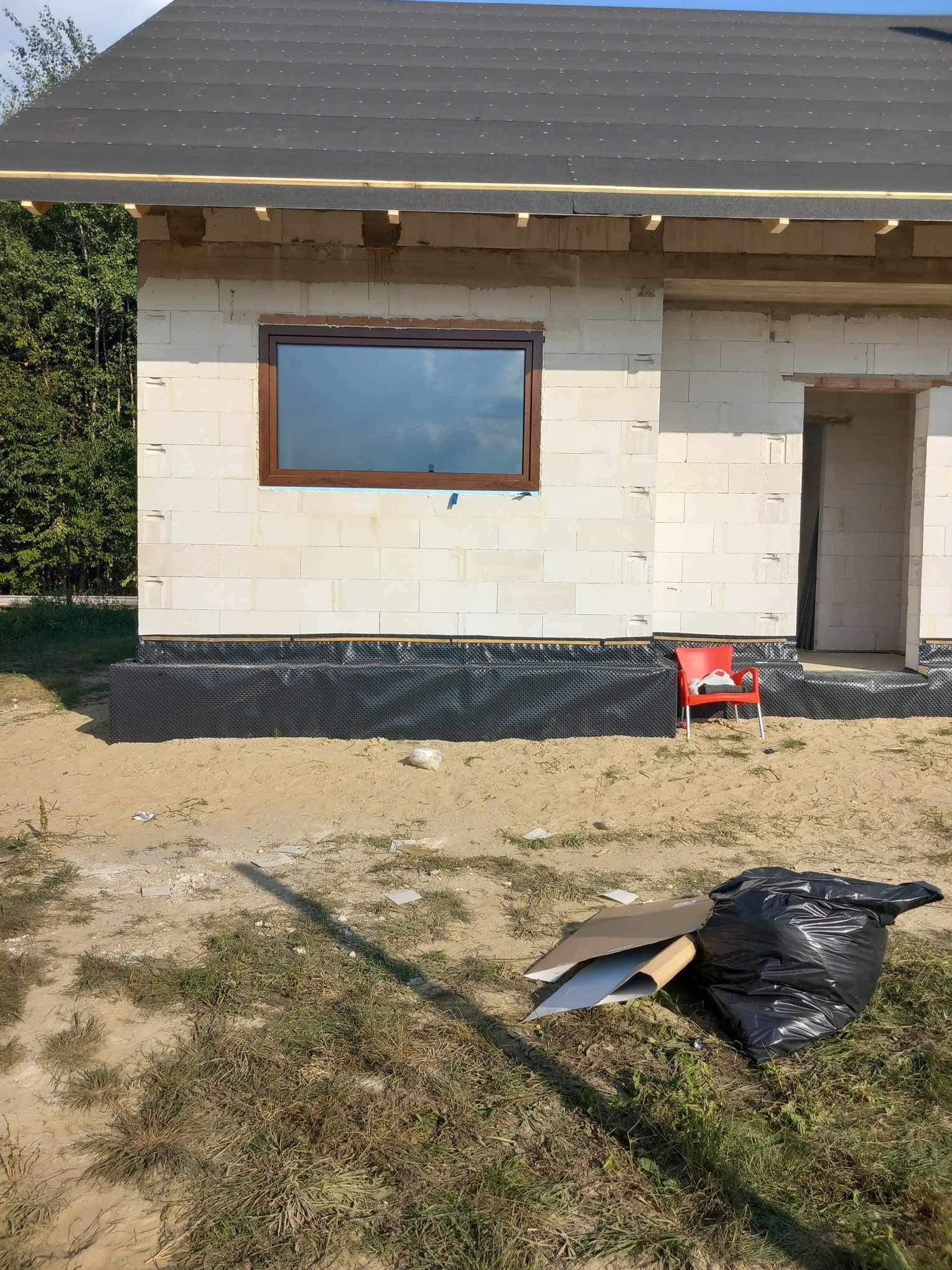 Surowy stan domu jednorodzinnego w trakcie budowy z zamontowanym oknem w brązowej ramie. Na pierwszym planie śmieci i gruz budowlany.