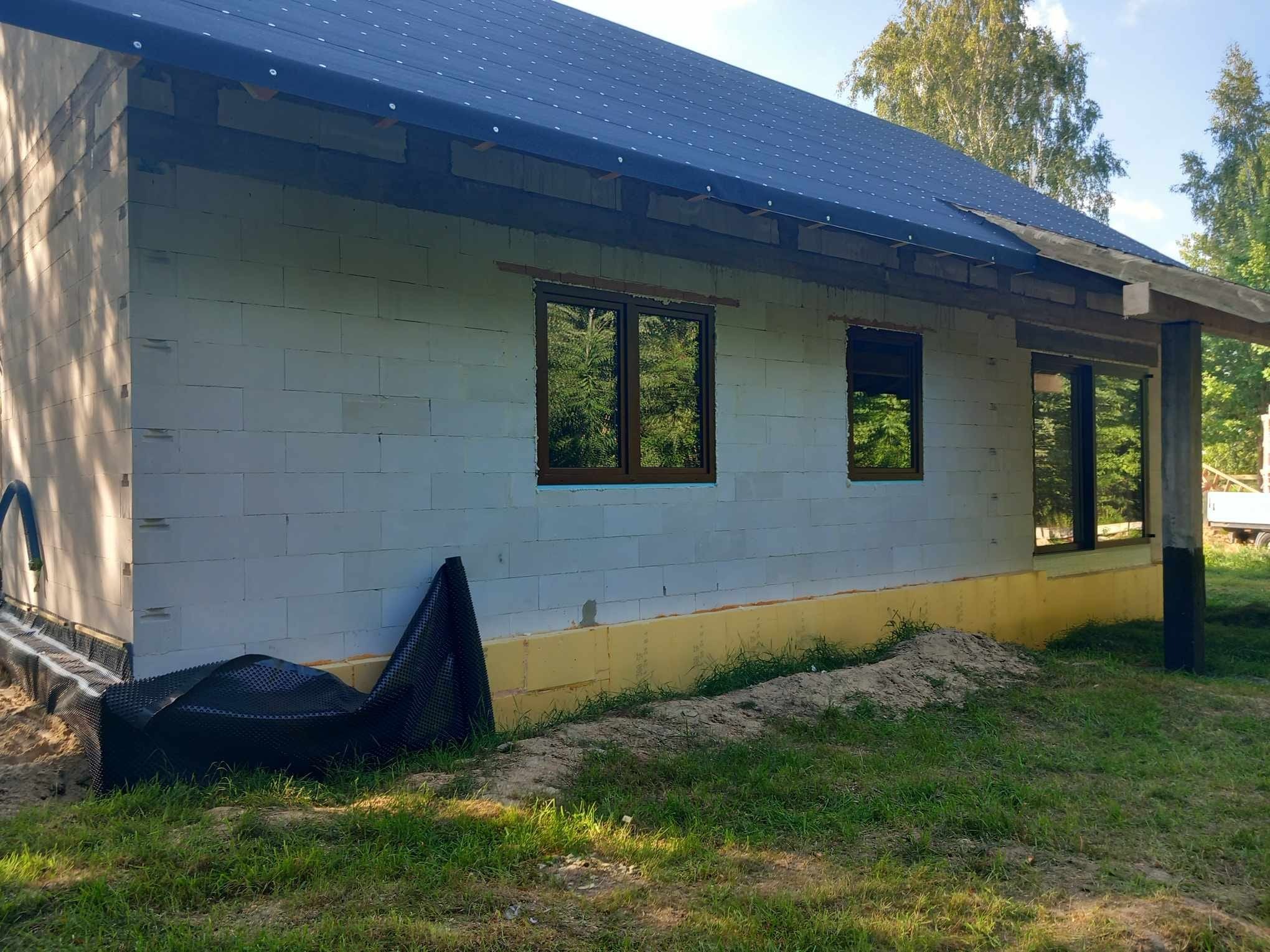 Budowa domu w trakcie: wstawione okna z brązowymi ramami, izolacja fundamentów i folia kubełkowa, ściany z bloczków, widok z zewnątrz w słoneczny dzień.