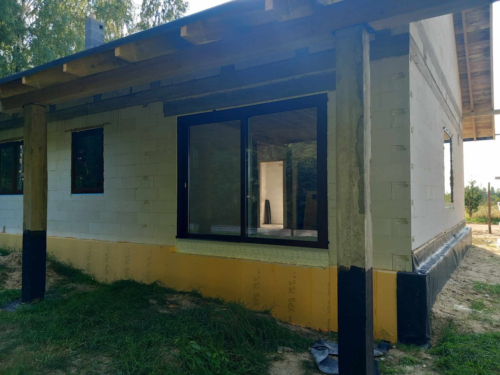 Surowy stan domu w budowie z zamontowanymi oknami z ciemnymi ramami, widoczne ocieplenie fundamentów. Dom z betonu komórkowego.