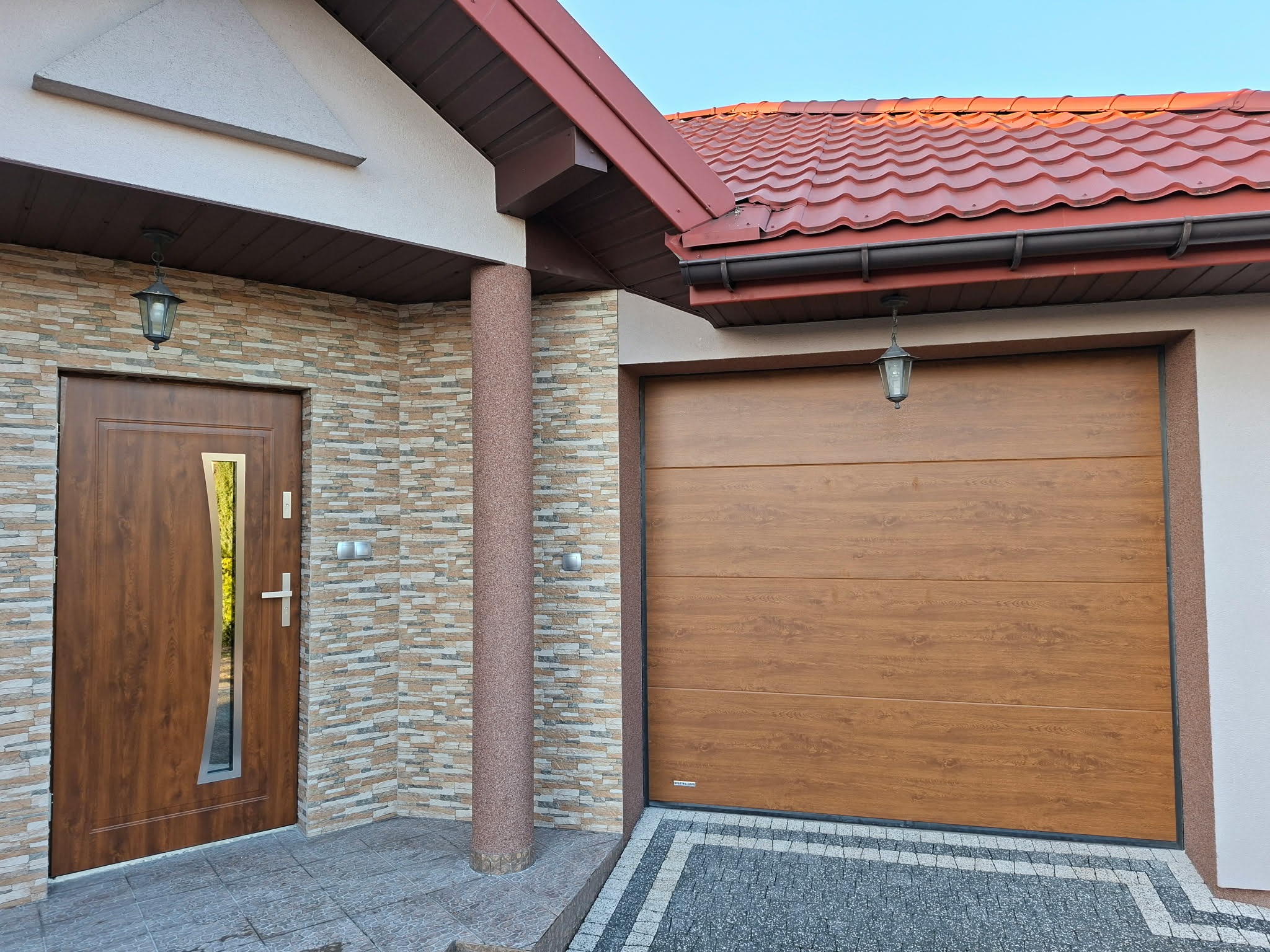 Brązowa brama garażowa segmentowa i drzwi wejściowe z szybą, imitujące drewno, zamontowane w elewacji z dekoracyjnej cegły, oświetlone lampami.