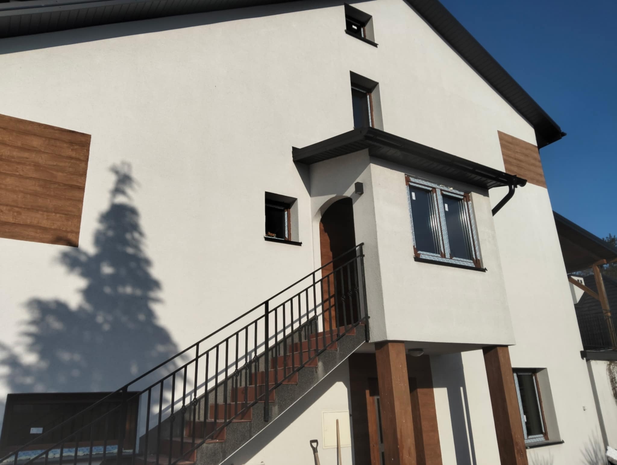 Nowoczesny dom z białymi ścianami i drewnianymi akcentami. Widoczne okna z folią ochronną, schody z czarną balustradą prowadzące do wejścia.
