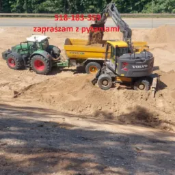 Żółta koparka Volvo nabiera ziemię do przyczepy ciągniętej przez zielony traktor na tle zielonych drzew. Widoczne numery telefonu.
