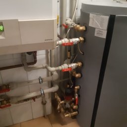 All-Fix - Biały piec gazowy Eurostar z podłączonymi rurami wodnymi i szary zbiornik akumulacyjny w kotłowni z beżową podłogą z płytek.