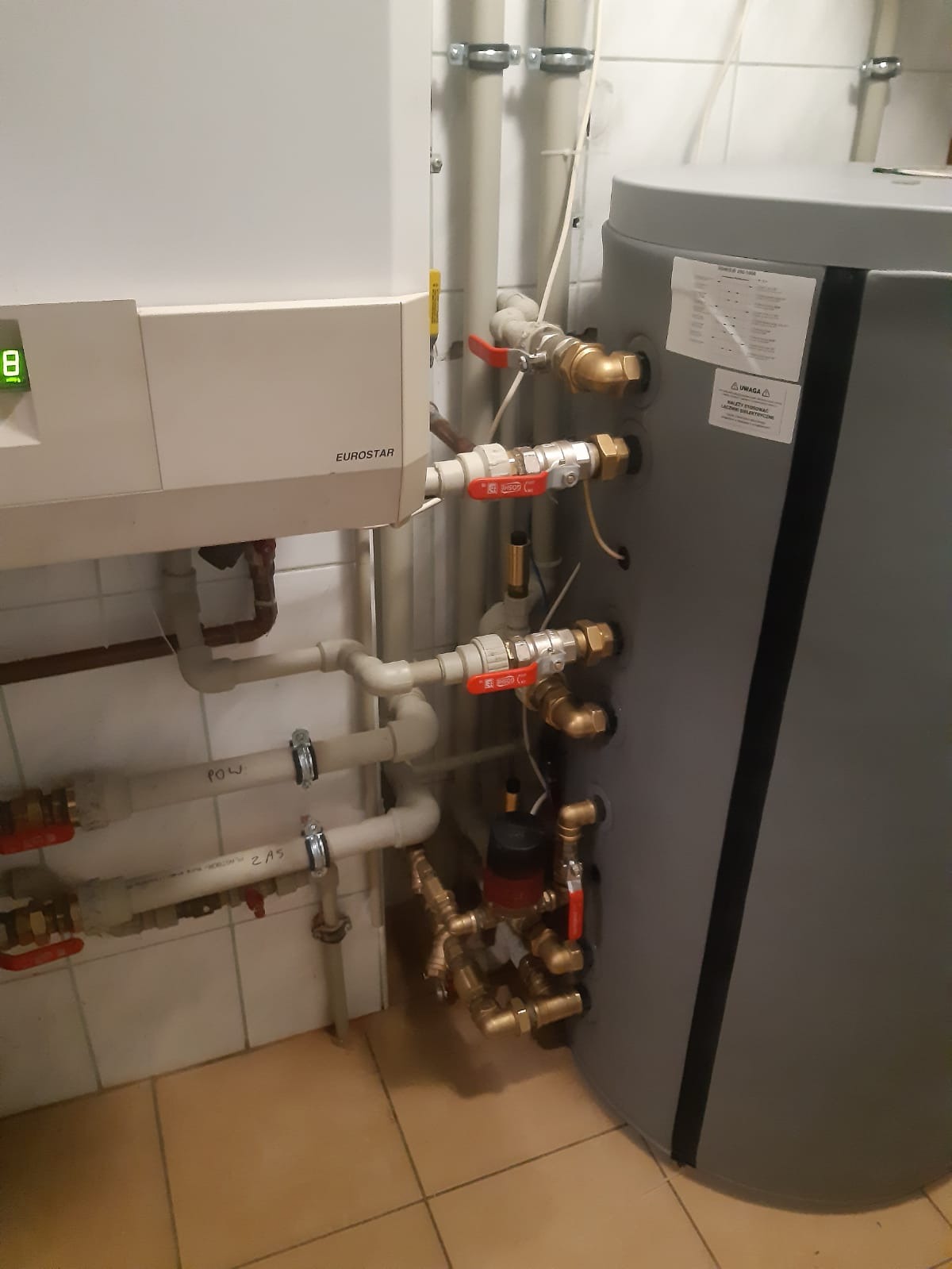 Biały piec gazowy Eurostar z podłączonymi rurami wodnymi i szary zbiornik akumulacyjny w kotłowni z beżową podłogą z płytek.