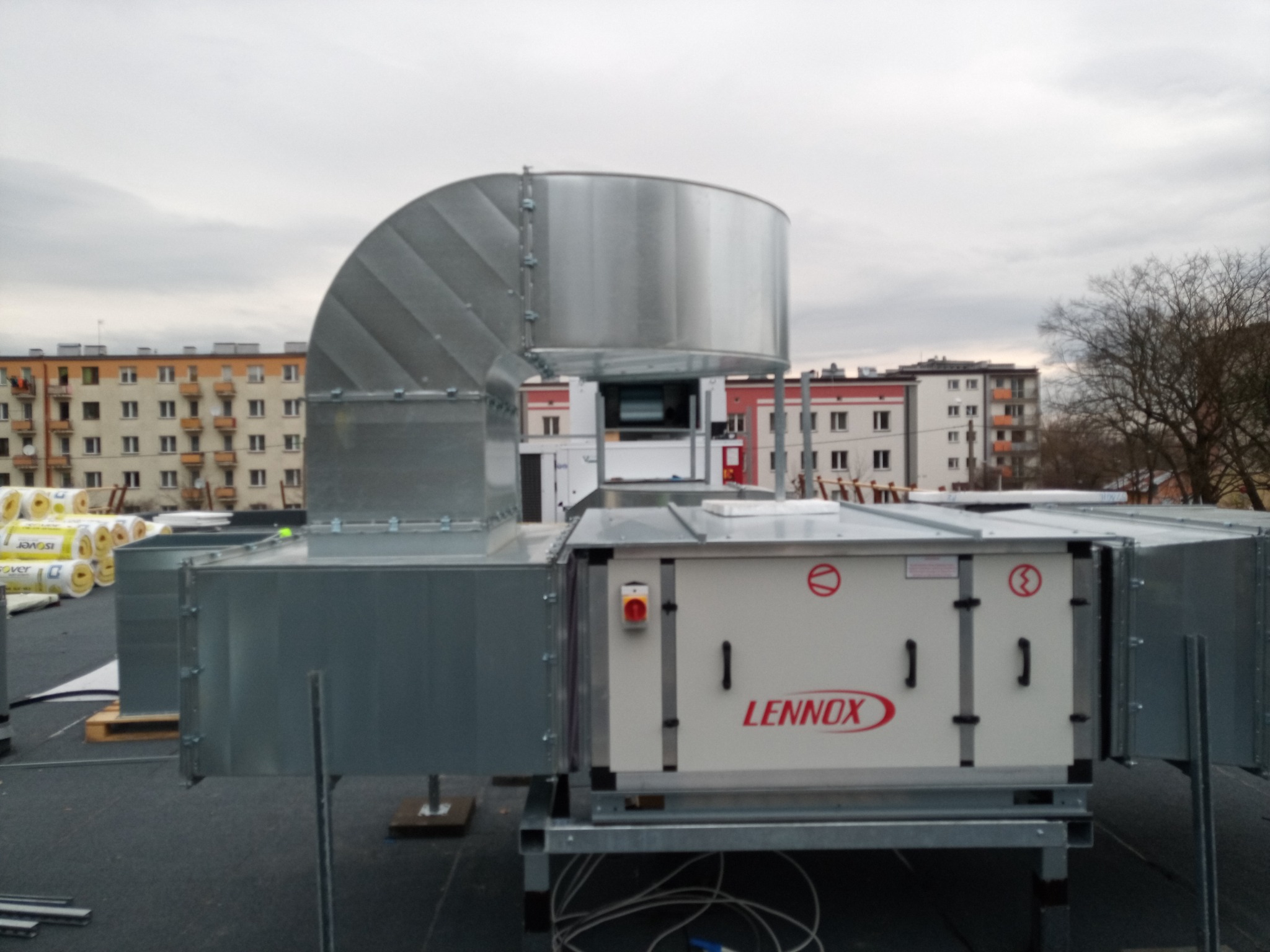 Szara centrala wentylacyjna Lennox z metalowymi kanałami na dachu budynku mieszkalnego, widoczne rolki izolacji.