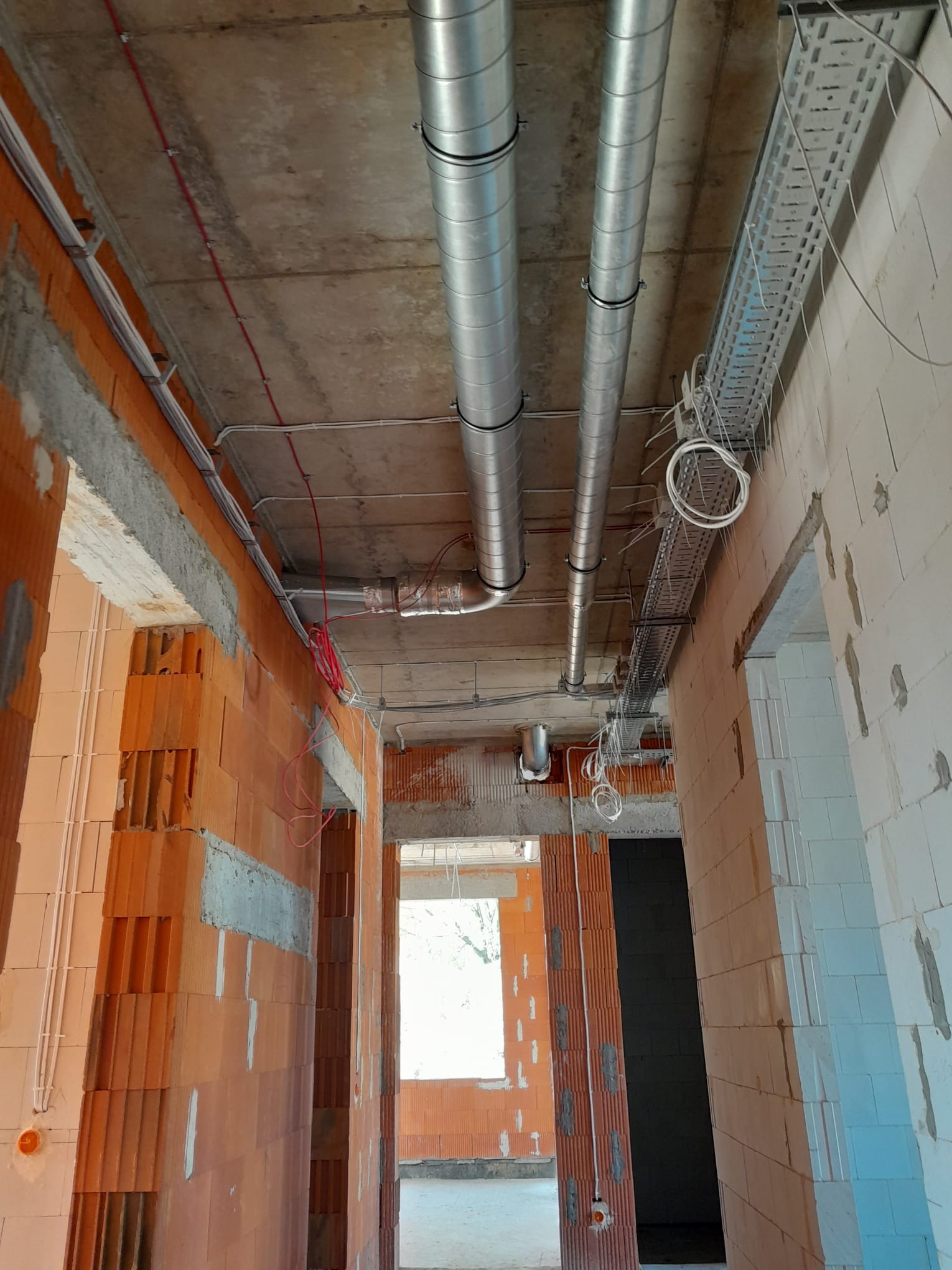 Wnętrze w trakcie montażu instalacji wentylacyjnej z widocznymi okrągłymi kanałami wentylacyjnymi pod sufitem, korytem kablowym, przewodami elektrycznymi i surowymi ścianami z cegły kratówki...