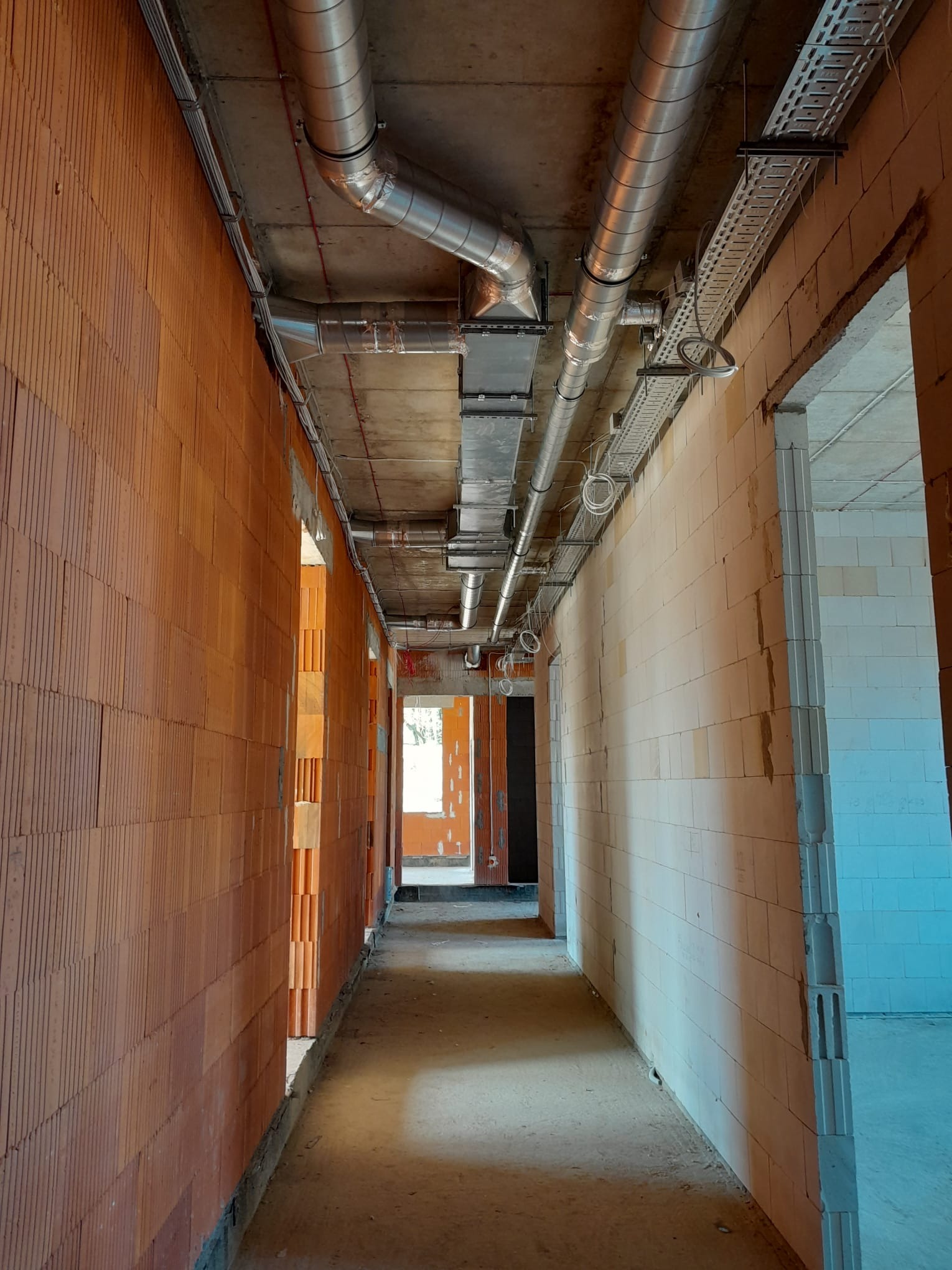 Wnętrze w trakcie budowy z widocznymi przewodami wentylacyjnymi pod sufitem, korytkami kablowymi i ścianami z cegły dziurawki.
