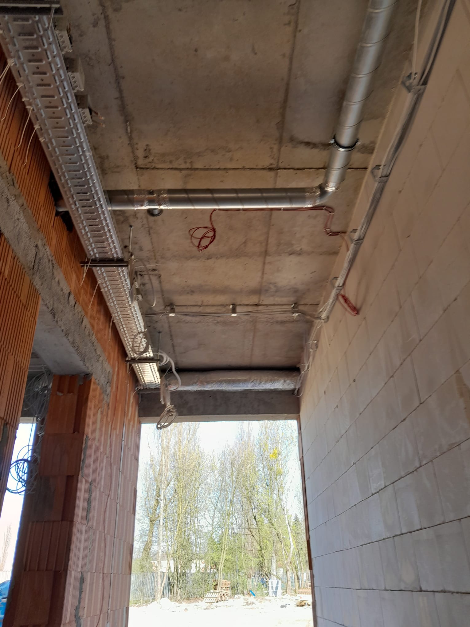 Surowy strop z widocznymi kanałami wentylacyjnymi, korytkami kablowymi i przewodami w stanie instalacji w budynku w trakcie budowy. Widoczne cegły i bloczki betonowe.