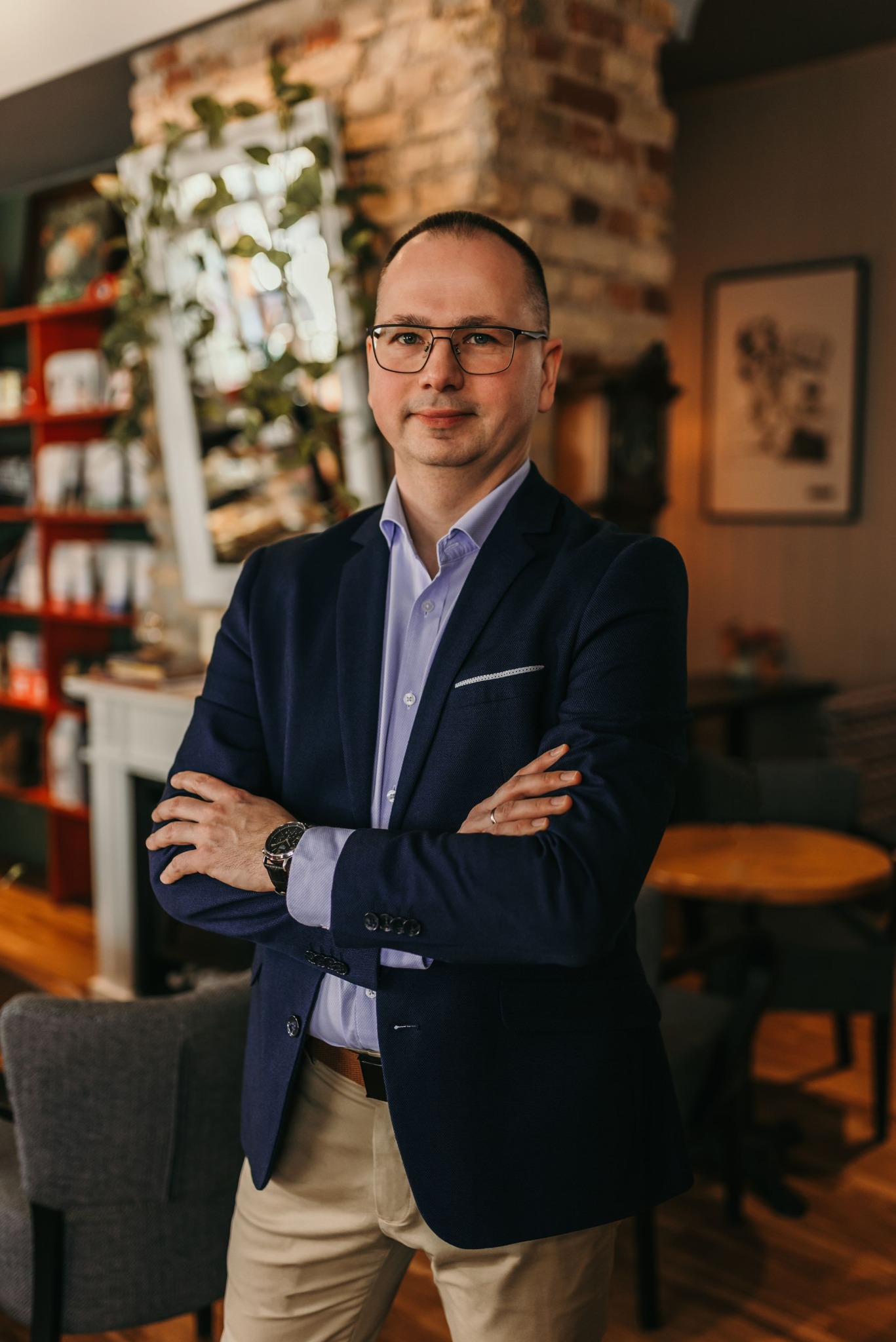 Mężczyzna w okularach, granatowej marynarce i beżowych spodniach, stoi z założonymi rękami na tle wnętrza kawiarni z ceglaną ścianą i regałem z książkami.