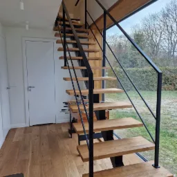 Nowoczesne schody z drewnianymi stopniami i czarną metalową balustradą, widok na ogród przez duże okno.