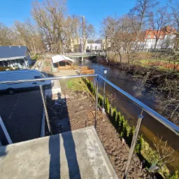 Błyszcząca, metalowa balustrada z widokiem na rzekę, drzewa i budynki w oddali, z zaparkowanym białym vanem w tle i cieniem dwóch osób w kadrze.