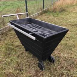 Czarny, metalowy grill na kółkach z uchwytami, stojący na trawie przed ogrodzeniem panelowym i drzewami.