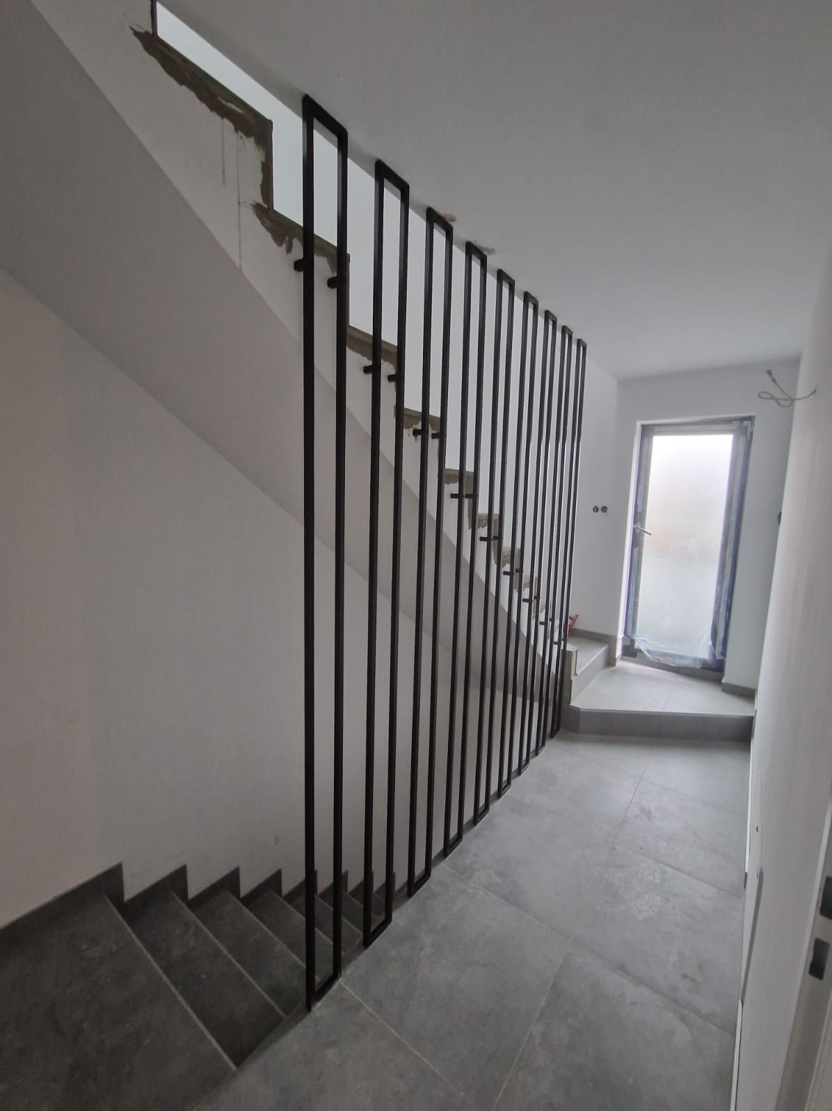 Minimalistyczna barierka schodowa z czarnych, pionowych profili stalowych, w surowym wnętrzu z szarymi schodami i podłogą, widoczne niedokończone wykończenia ścian.