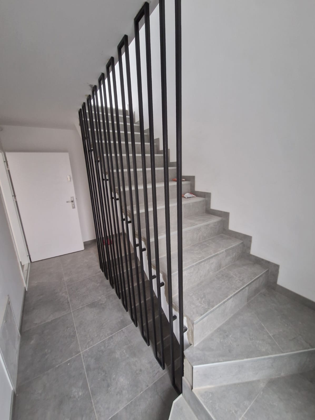 Schody wyłożone szarymi płytkami z pionową, czarną balustradą z prostokątnych profili, widok z dołu na białe drzwi.