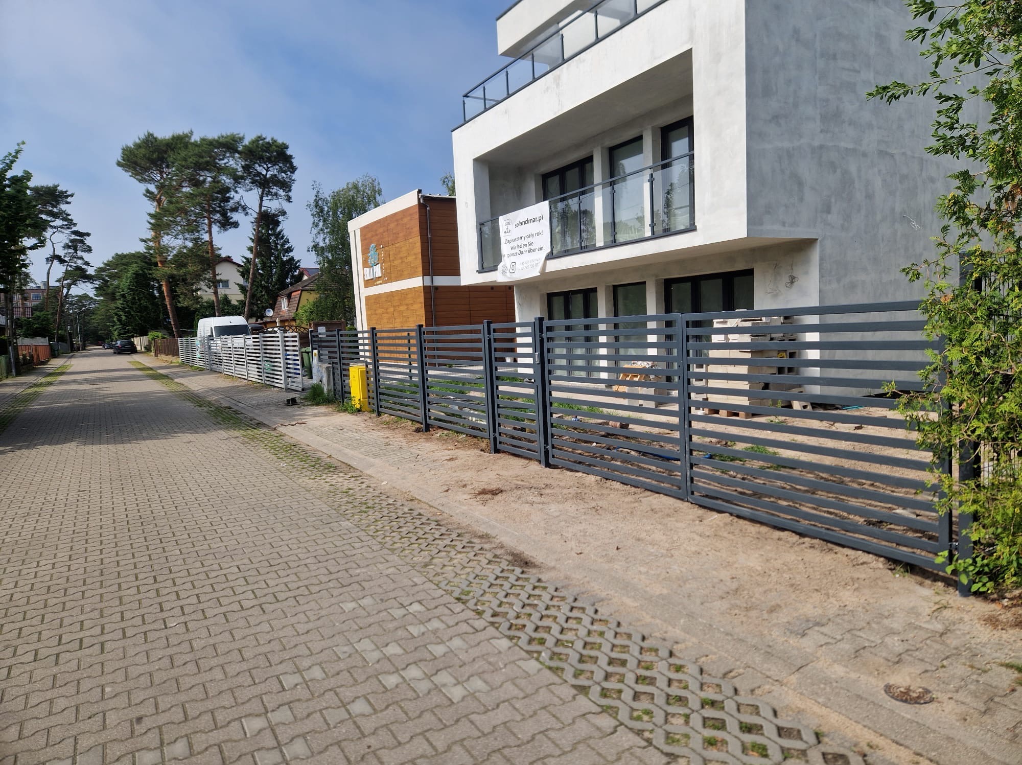 Nowoczesne ogrodzenie z poziomych paneli w kolorze grafitowym, przylegające do nowo wybudowanego budynku z balkonami i szklanymi balustradami, widok z brukowanej ulicy.