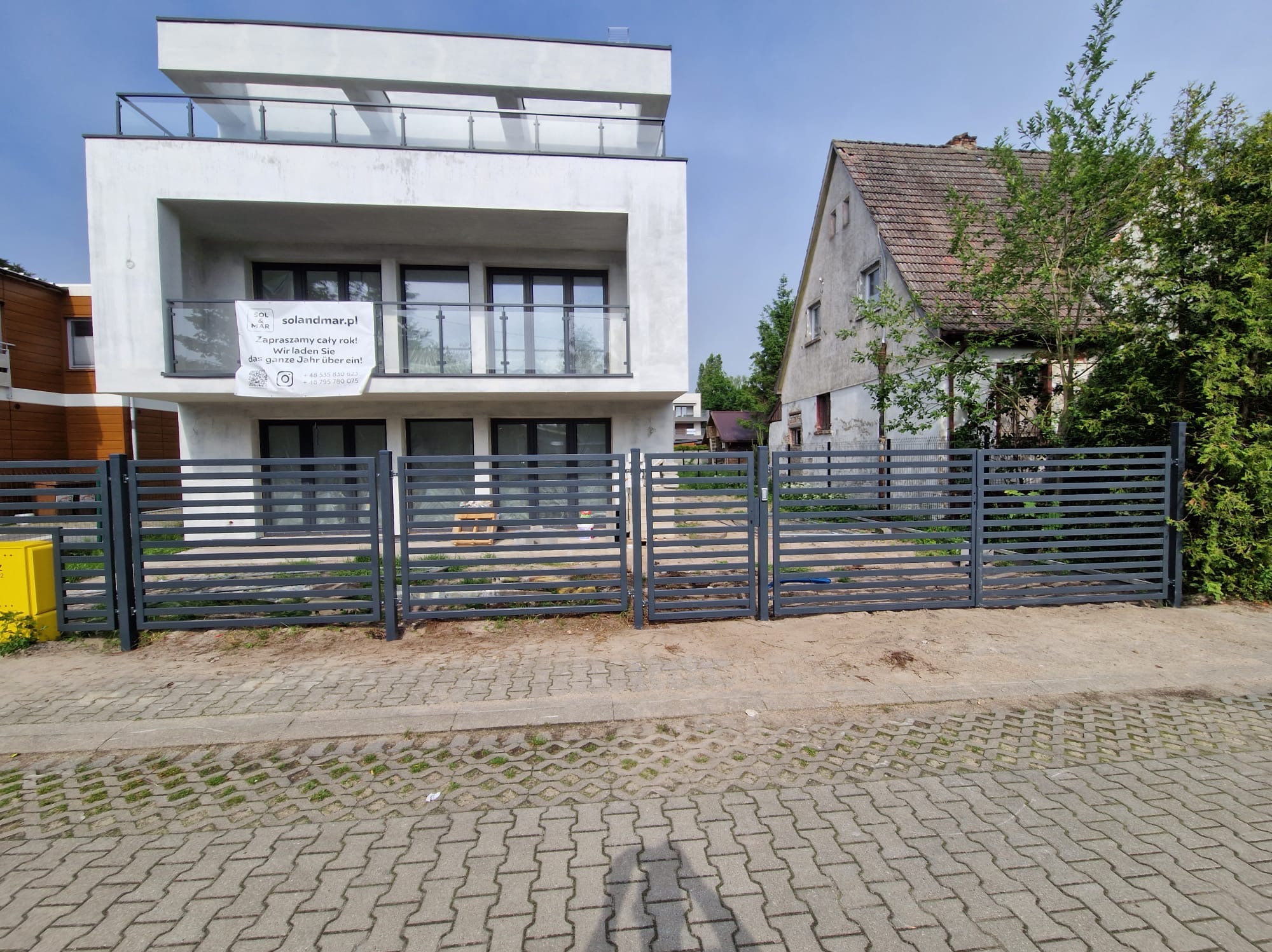 Nowoczesne, poziome, szare ogrodzenie panelowe przed nowym, białym domem z balkonami i reklamą 'solandmar.pl', w tle starszy dom z czerwonej cegły.
