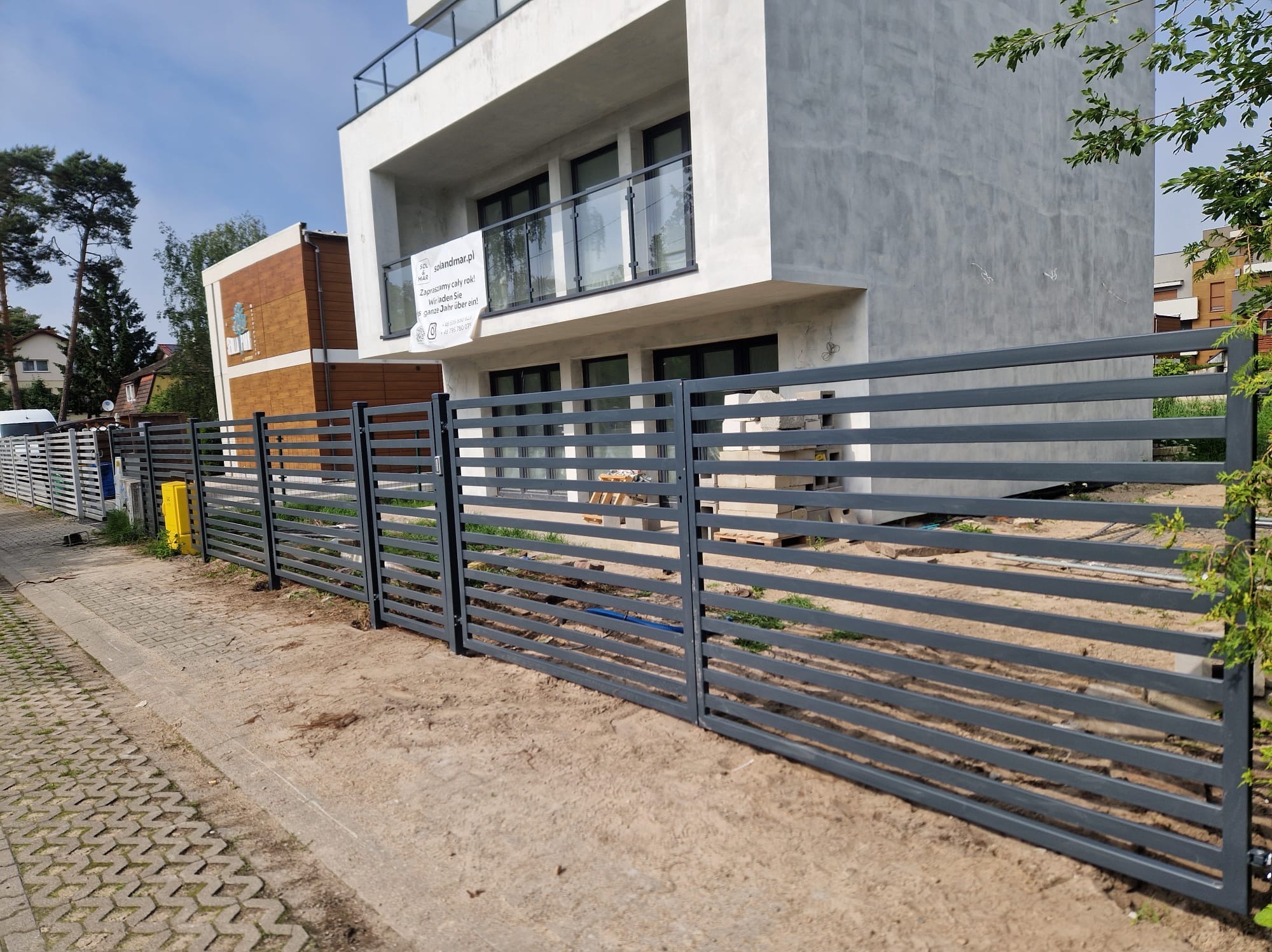 Nowoczesne ogrodzenie panelowe z poziomych, szarych profili, na tle budynku z balkonem i reklamą solandmar.pl.