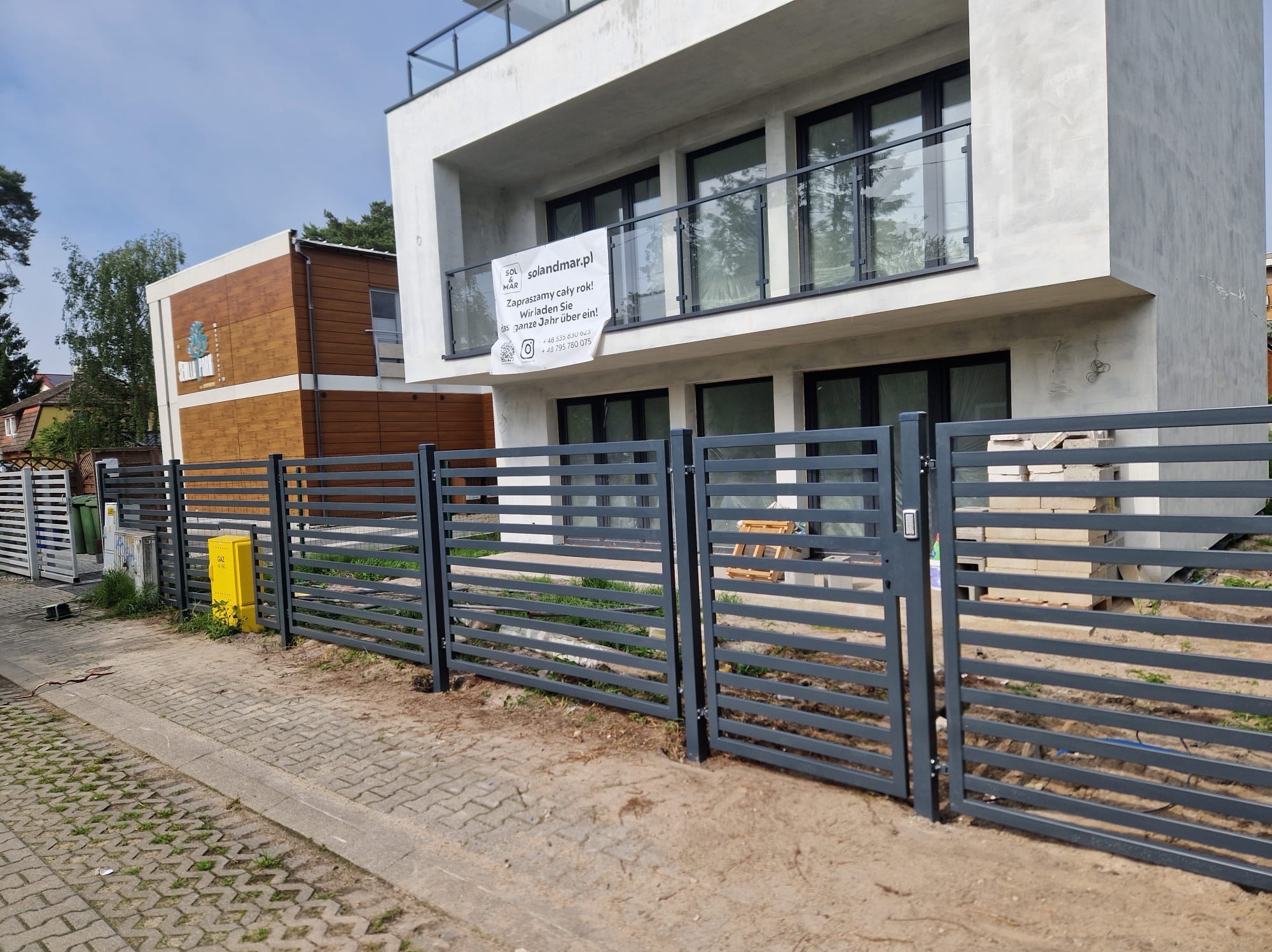 Nowoczesne, poziome ogrodzenie panelowe w kolorze grafitowym przed dwukondygnacyjnym domem z białym tynkiem i szklaną balustradą balkonową; widoczna brama wjazdowa z domofonem oraz skrzynka gazowa.