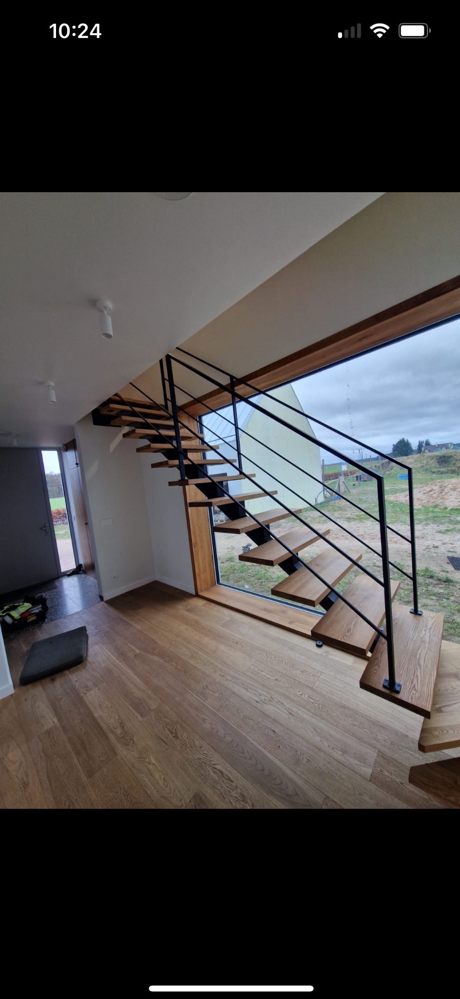 Nowoczesne schody z drewnianymi stopniami i czarną metalową balustradą, zamontowane w domu w Zaleszczycach, widoczne przez duże okno z widokiem na zewnątrz.