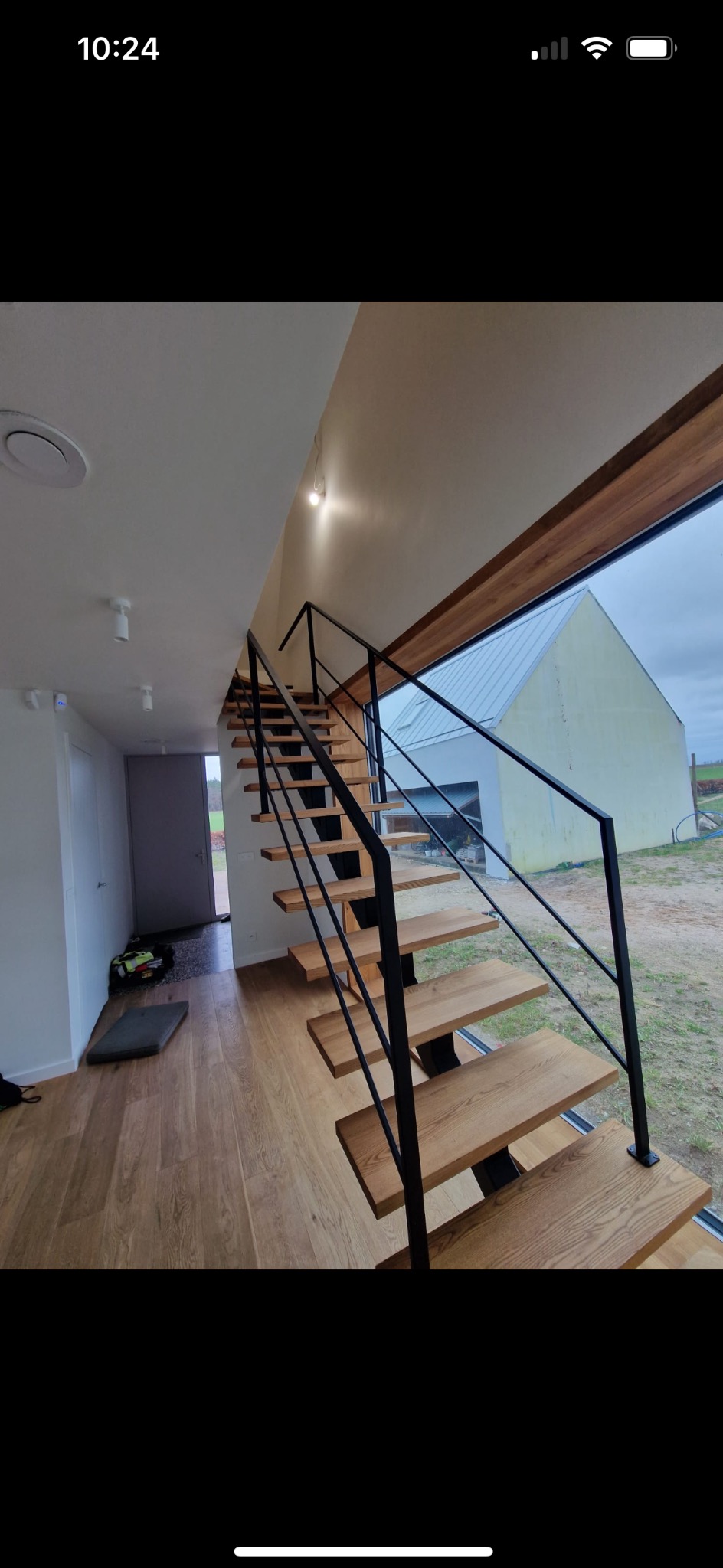 Wnętrze domu z drewnianymi schodami o konstrukcji wspornikowej i czarną, minimalistyczną balustradą, widok przez okno na budynek gospodarczy.