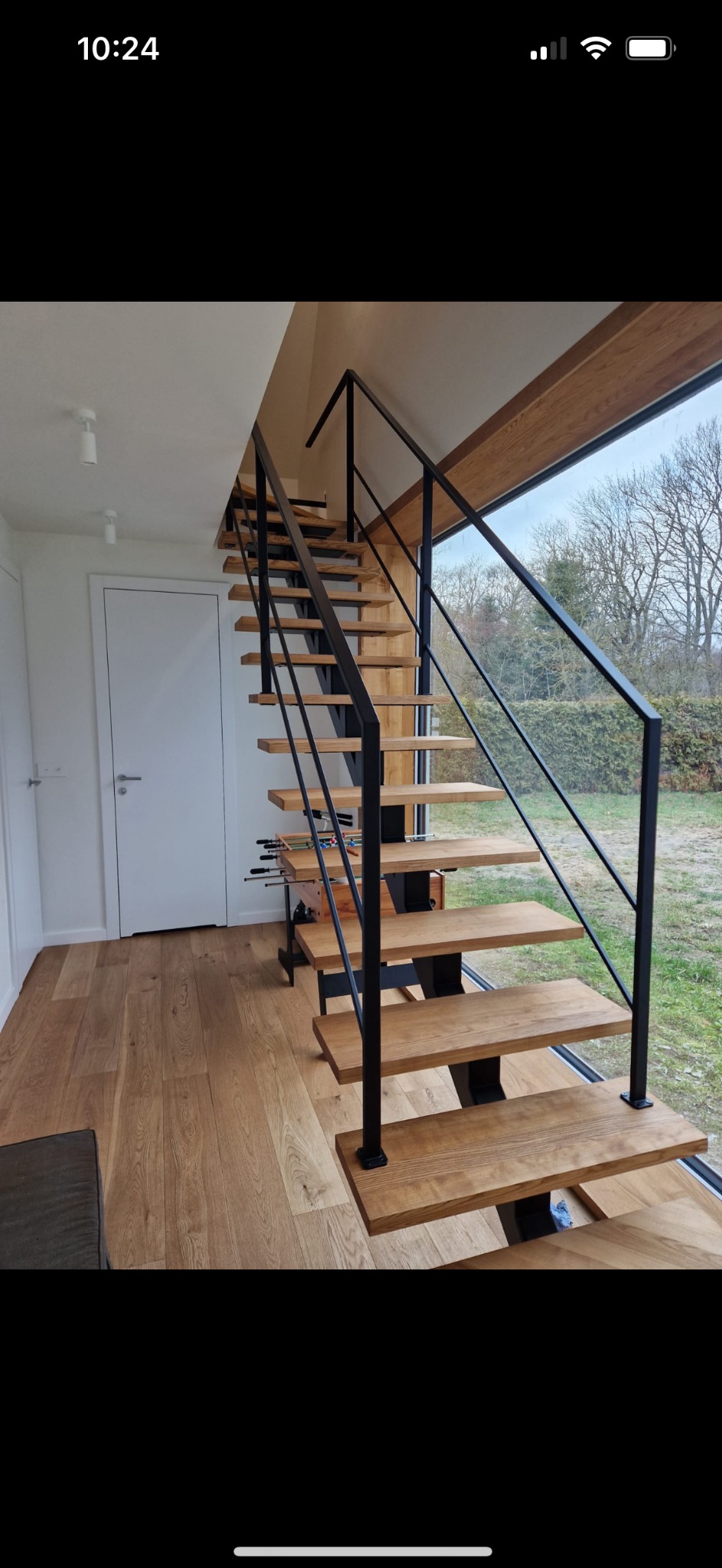 Nowoczesne schody z drewnianymi stopniami i czarną metalową balustradą, widok na ogród przez duże okno.
