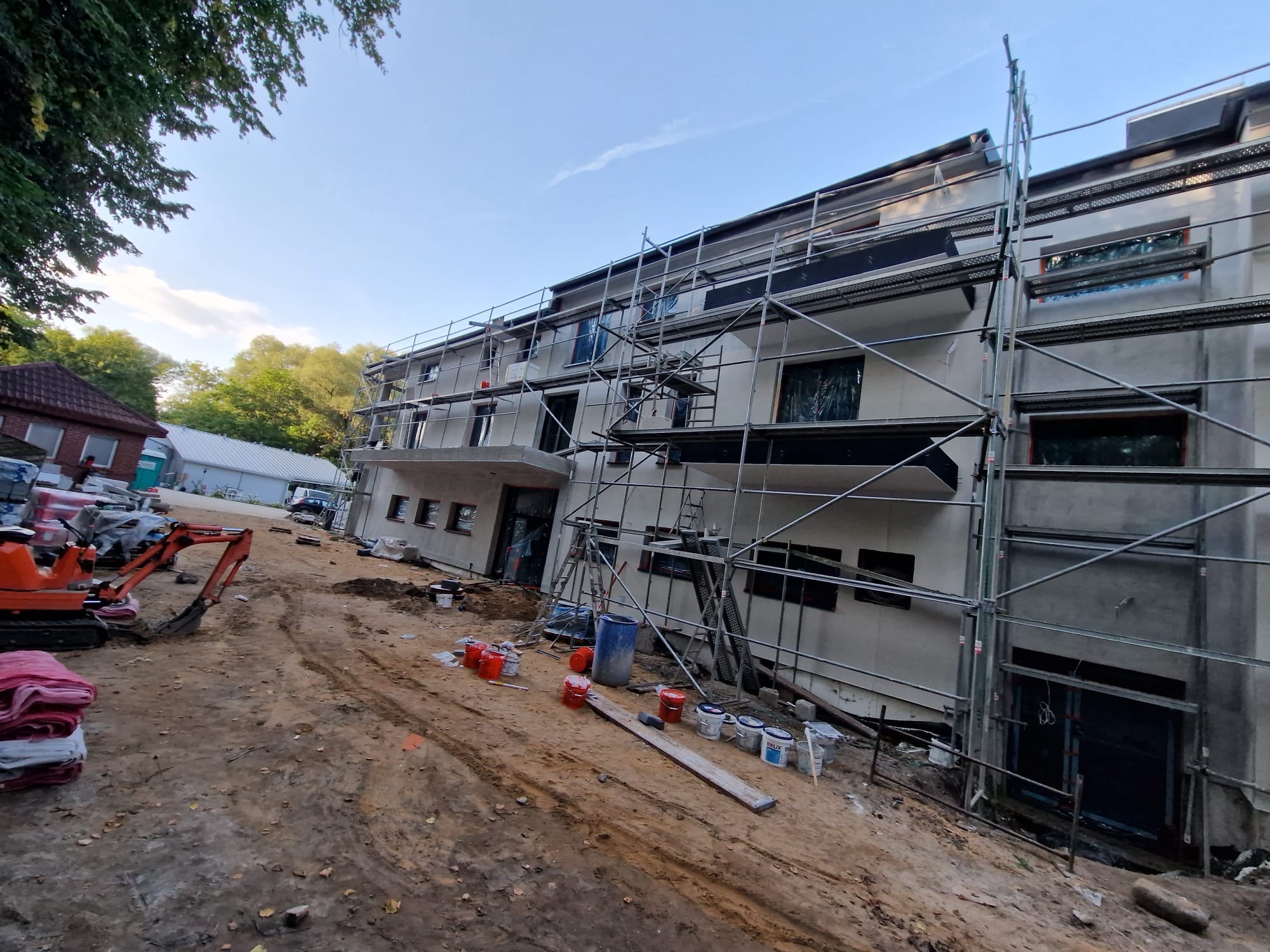 Budynek w trakcie prac wykończeniowych, z rusztowaniem, farbami i mini koparką na placu budowy.