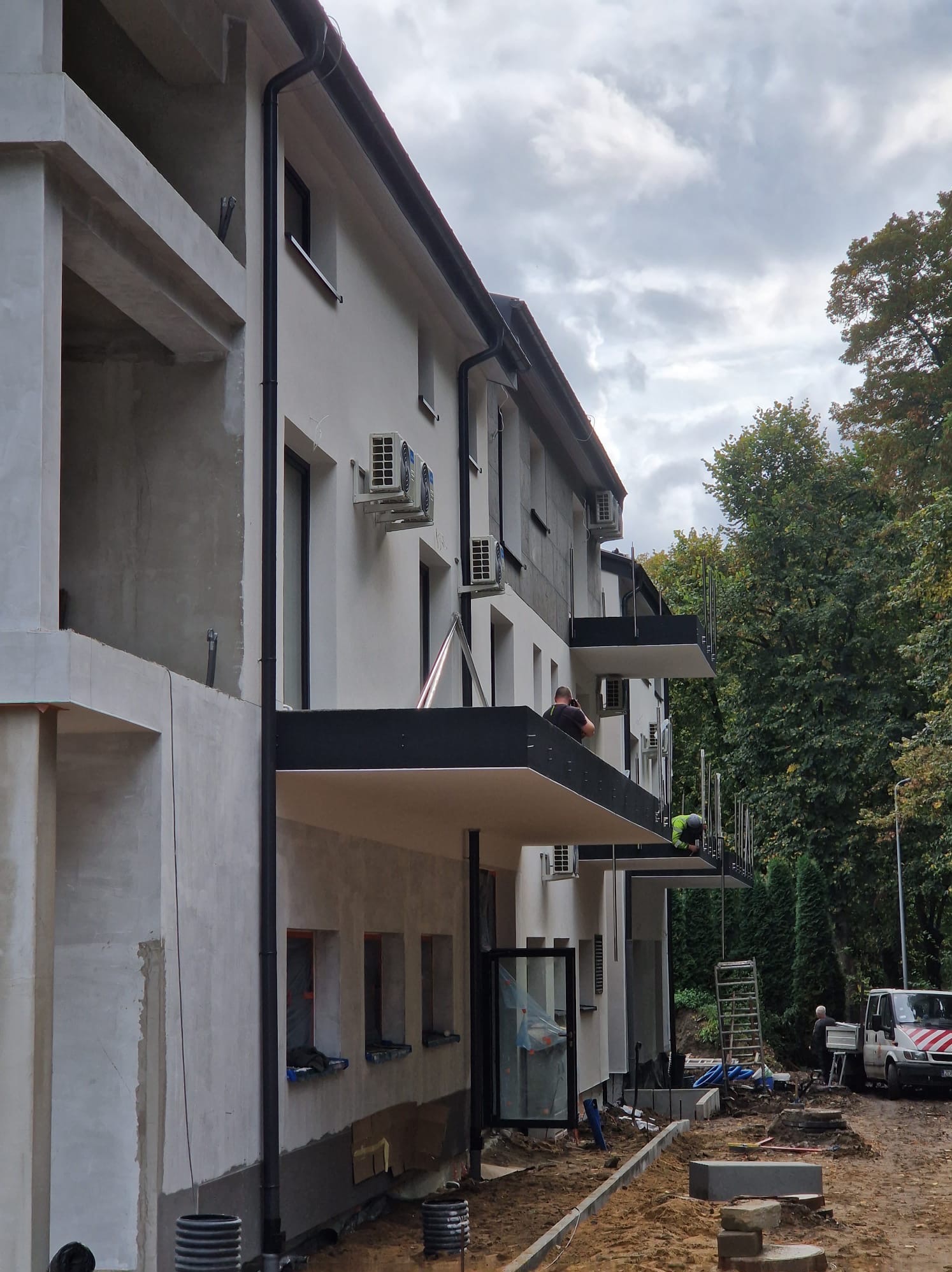 Budynek wielorodzinny w trakcie wykańczania z zamontowanymi balkonami z czarną podłogą i srebrnymi barierkami, dwóch pracowników montuje barierki, na elewacji widoczne klimatyzatory, teren wokół...
