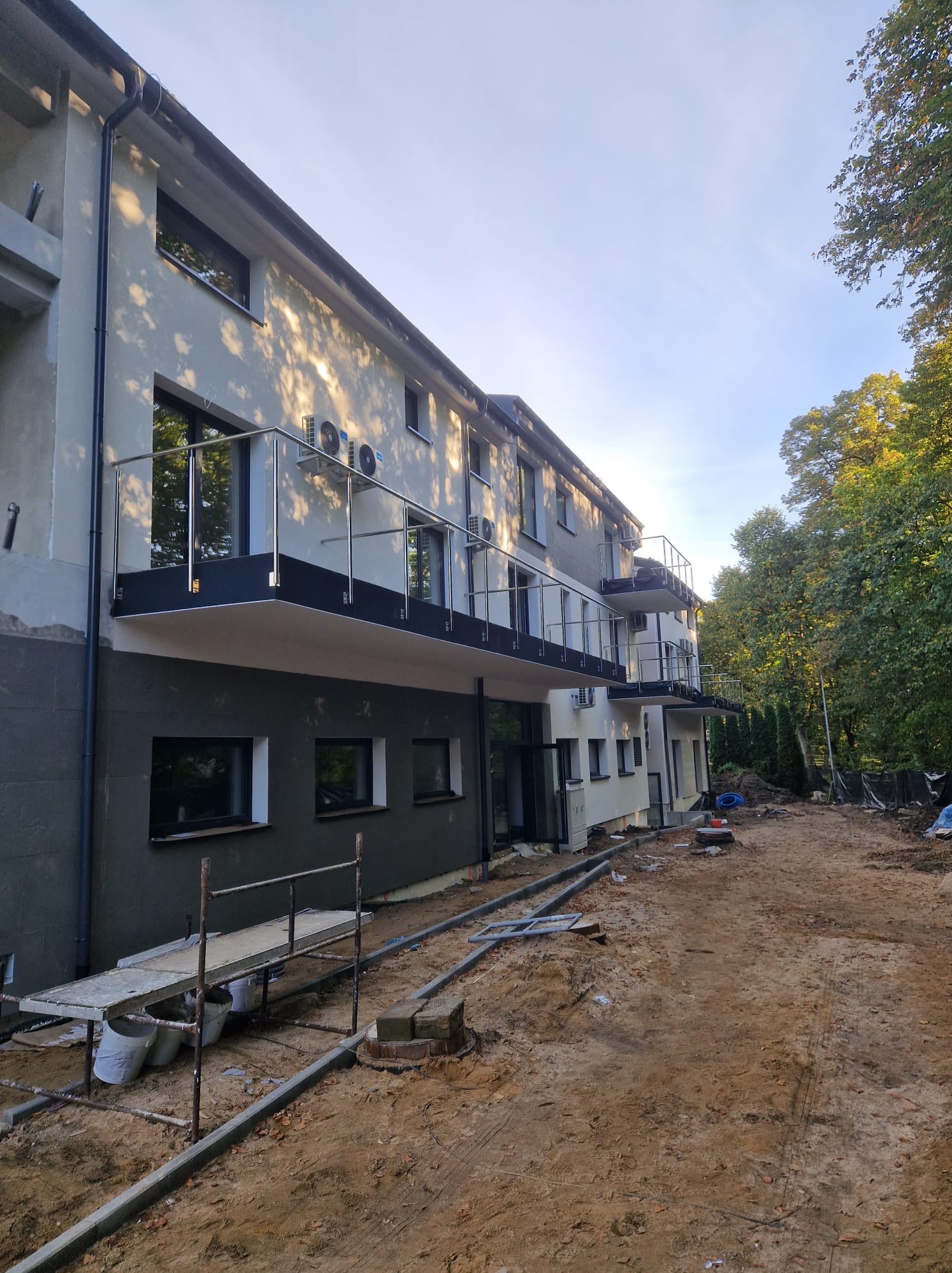 Budynek w trakcie wykończenia z balkonami z metalowymi balustradami, widoczne rusztowania i teren budowy.