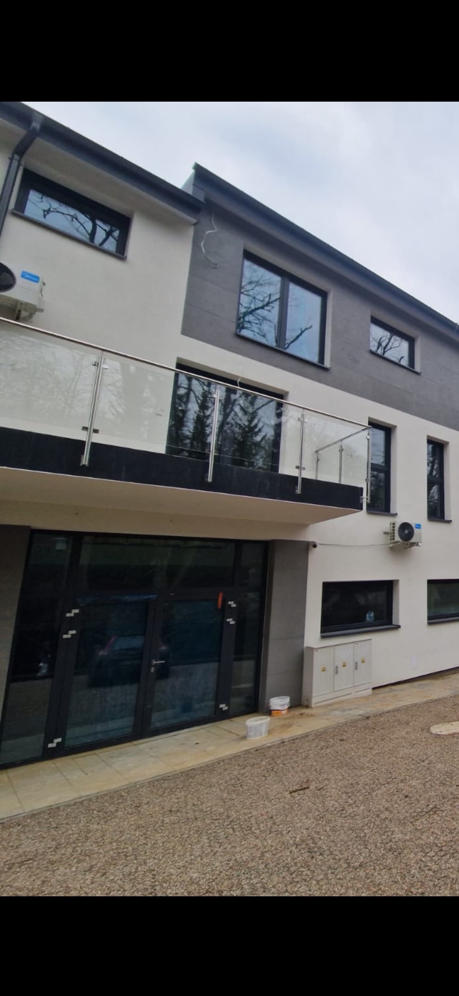 Nowoczesny dwupiętrowy budynek z balkonem zabezpieczonym przeszkloną balustradą z metalowymi słupkami, widoczne okna, drzwi wejściowe i klimatyzatory.