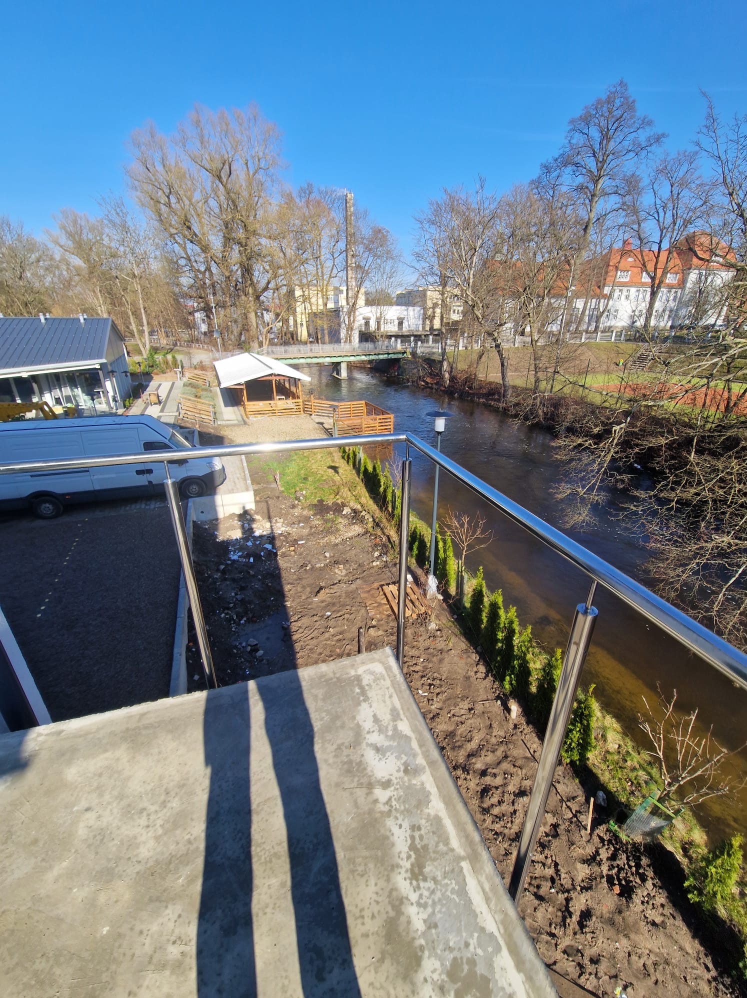 Błyszcząca, metalowa balustrada z widokiem na rzekę, drzewa i budynki w oddali, z zaparkowanym białym vanem w tle i cieniem dwóch osób w kadrze.