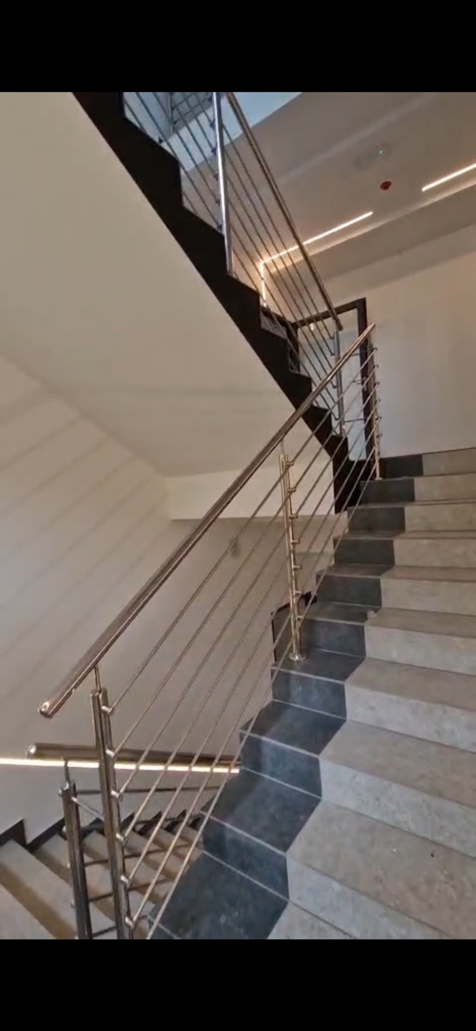 Wewnętrzne schody z szarymi stopniami i nowoczesną balustradą ze stali nierdzewnej z poziomymi prętami, oświetlenie LED na suficie.