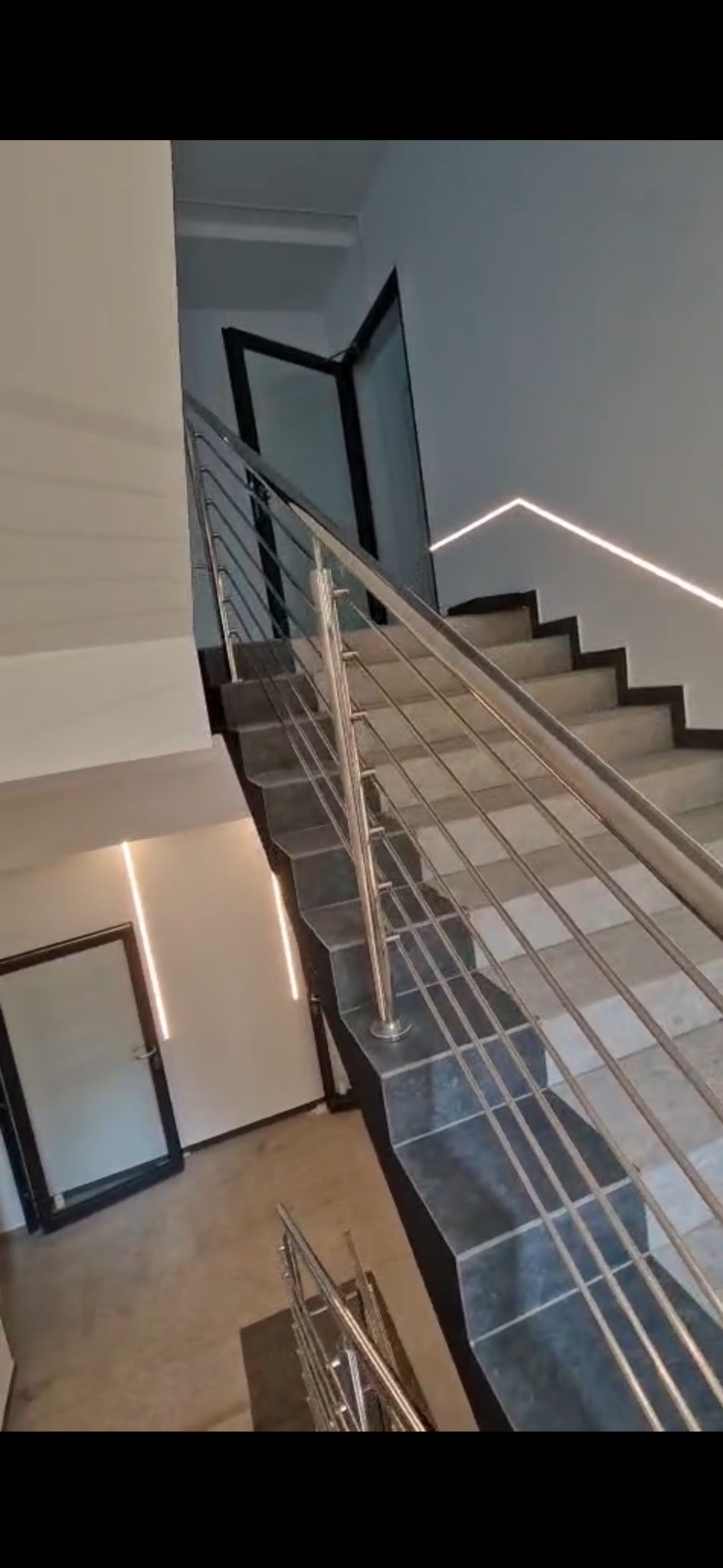 Schody wykończone ciemnymi płytkami z balustradą ze stali nierdzewnej, oświetlenie LED wzdłuż ściany i nad otwartymi drzwiami.