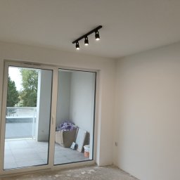 TOM-BUD - Jasne wnętrze z dużym oknem balkonowym i nowoczesnym oświetleniem sufitowym. Ściany świeżo pomalowane na biało, podłoga przygotowana do dalszych prac wykończeniowych.