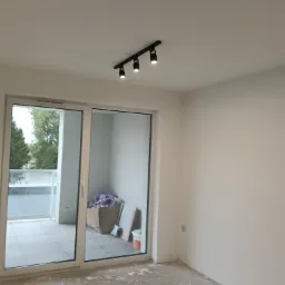 Jasne wnętrze z dużym oknem balkonowym i nowoczesnym oświetleniem sufitowym. Ściany świeżo pomalowane na biało, podłoga przygotowana do dalszych prac wykończeniowych.