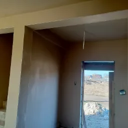 Świeżo nałożona gładź szpachlowa na ścianach i suficie w surowym wnętrzu, widoczne przejście do innego pomieszczenia i drzwi na zewnątrz.