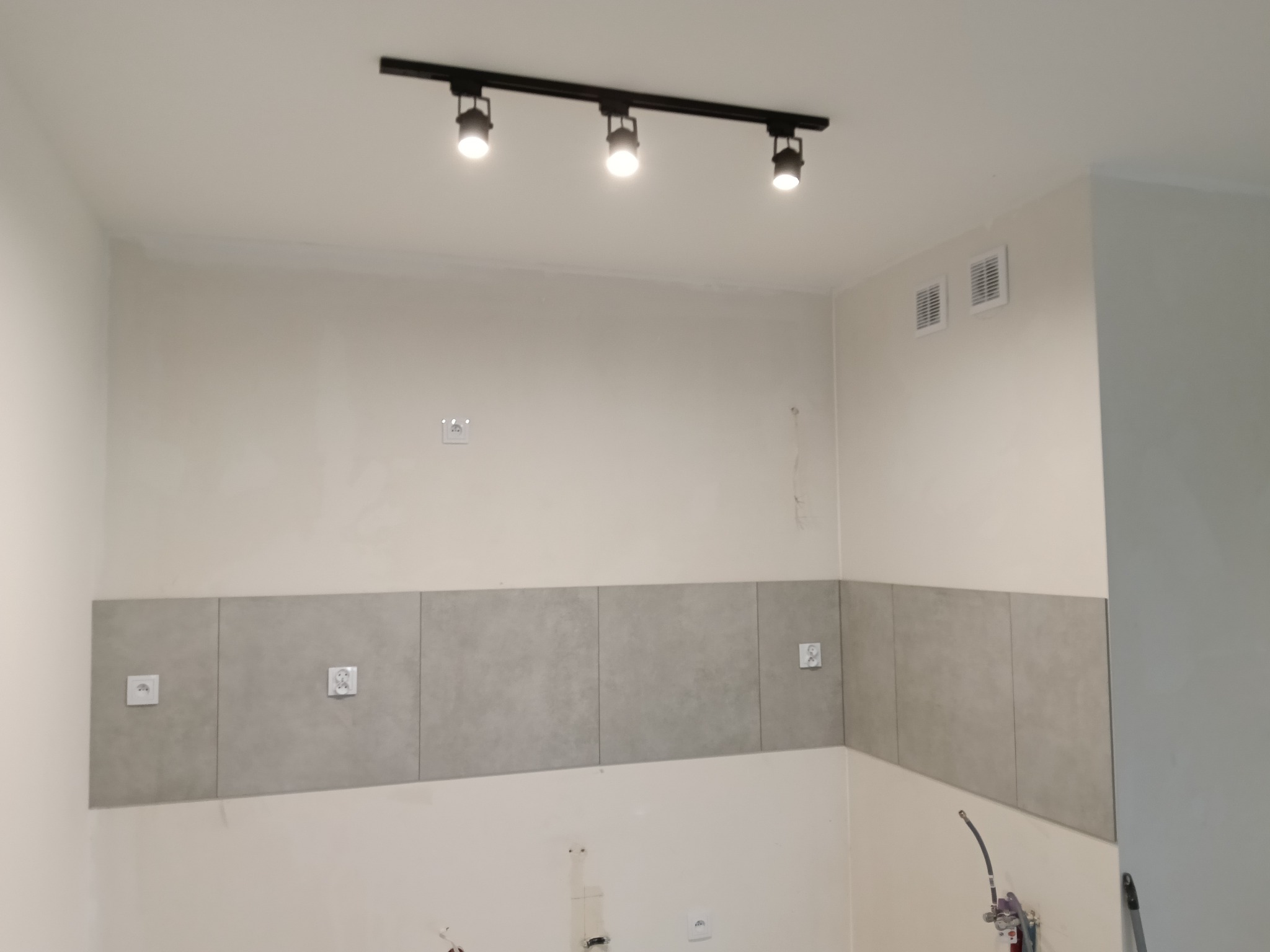 Fragment wnętrza w trakcie prac: szare płytki częściowo ułożone na ścianie, widoczne gniazdka elektryczne, lampa sufitowa i ślady po szpachlowaniu.